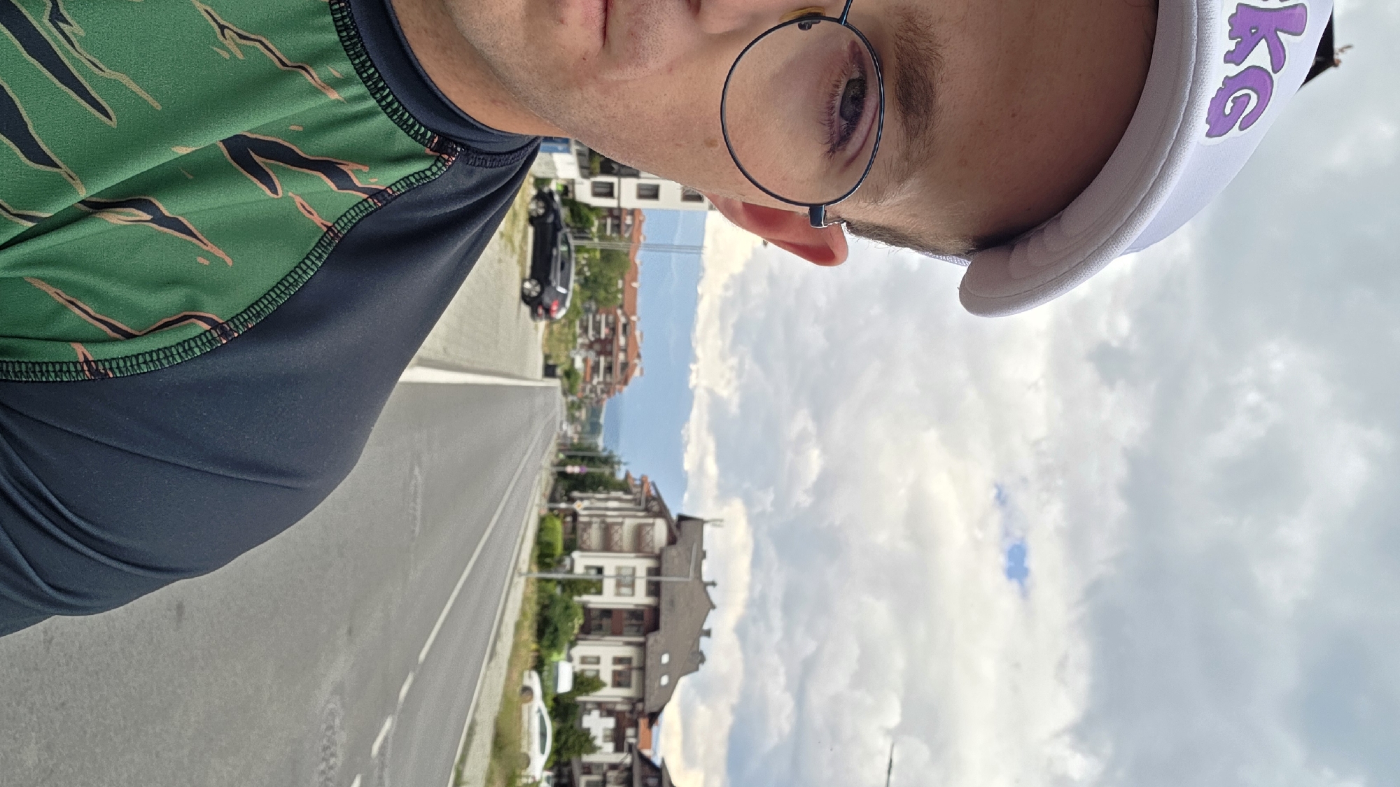 This is a first-person, upside-down selfie taken outdoors. The subject is a person wearing a green and black patterned t-shirt, a white visor with purple lettering that appears to say "KG", and round-rimmed glasses. Only the top portion of the person's face is visible, with their eye looking towards the camera.
The background shows a street with a road and a sidewalk, lined with multi-story buildings with reddish-brown roofs and balconies. Trees and greenery are interspersed between the buildings. A black car is parked on the side of the road, and another white car is partially visible further down. The sky is cloudy with patches of blue.
The image was likely taken during the daytime, with the cloudy sky suggesting potentially overcast or variable weather conditions. The architecture of the buildings and the overall scene suggest a European setting, specifically Bansko, Bulgaria, as indicated by the location context. The selfie style and focus on the face, combined with the outdoor setting, imply the person is perhaps on a walk, run, or enjoying an activity outdoors. There are no strong emotional cues conveyed due to the limited view of the subject's face.