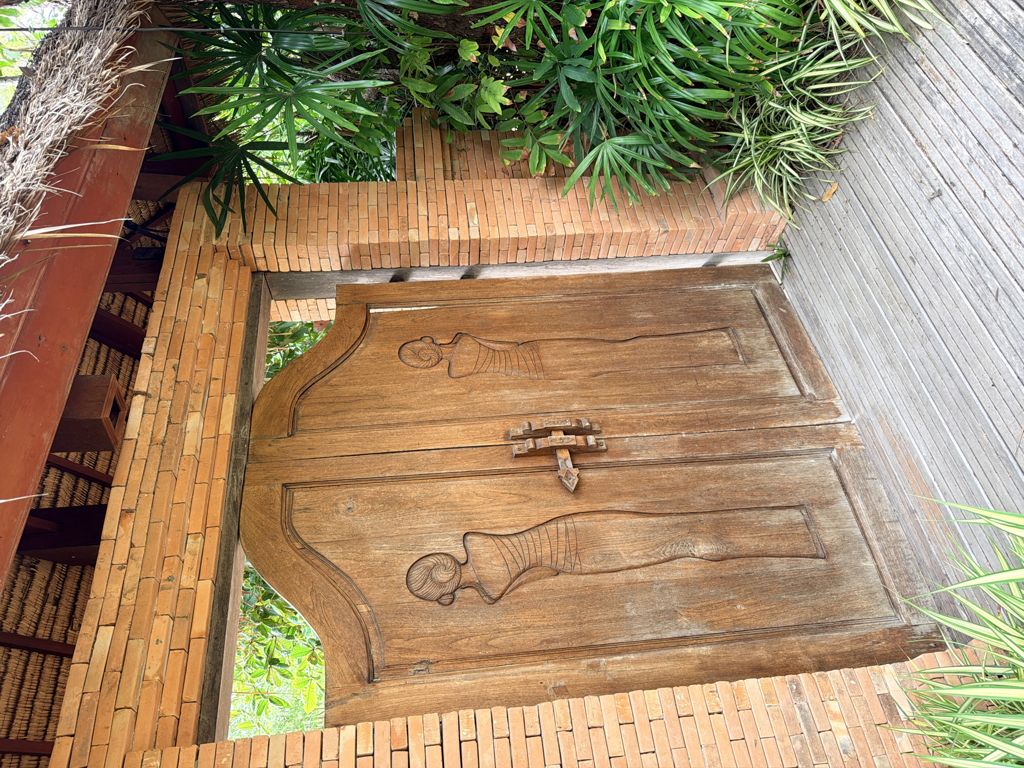 In Ko Samui District, Thailand, a weathered wooden double door, intricately carved with the figures of two women in traditional attire, stands as the focal point of this scene. The doors are set within a brick archway, constructed from light orange bricks. Above the archway, a thatched roof with exposed wooden beams is visible. To the right of the doors, lush green palm fronds and other tropical foliage spill over the brickwork, creating a vibrant contrast with the earthy tones of the architecture. The ground in front of the doors is paved with a wooden walkway, its planks running horizontally. The overall impression is one of natural beauty integrated with traditional craftsmanship, suggestive of a resort or a tastefully designed residential entrance.
