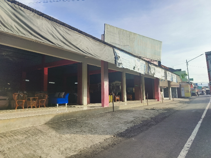 The image shows a street scene in Cikalong, Indonesia, located at the coordinates -6.733 latitude and 107.44 longitude. It appears to be a commercial area with several storefronts on the left side of the image. The storefronts are open to the street, and there are a few pieces of furniture, including wooden chairs and a blue couch, displayed in front of the building. The street itself is paved and appears to be relatively empty. The buildings have a weathered appearance with a combination of gray, pink, and green paint. One building has a large banner that is faded and torn. There are no people visible in the image. The sky is clear and blue. The street is slightly sloped, and the ground in front of the building is rough and uneven. The image captures a moment of quiet in a seemingly ordinary commercial area of Cikalong.