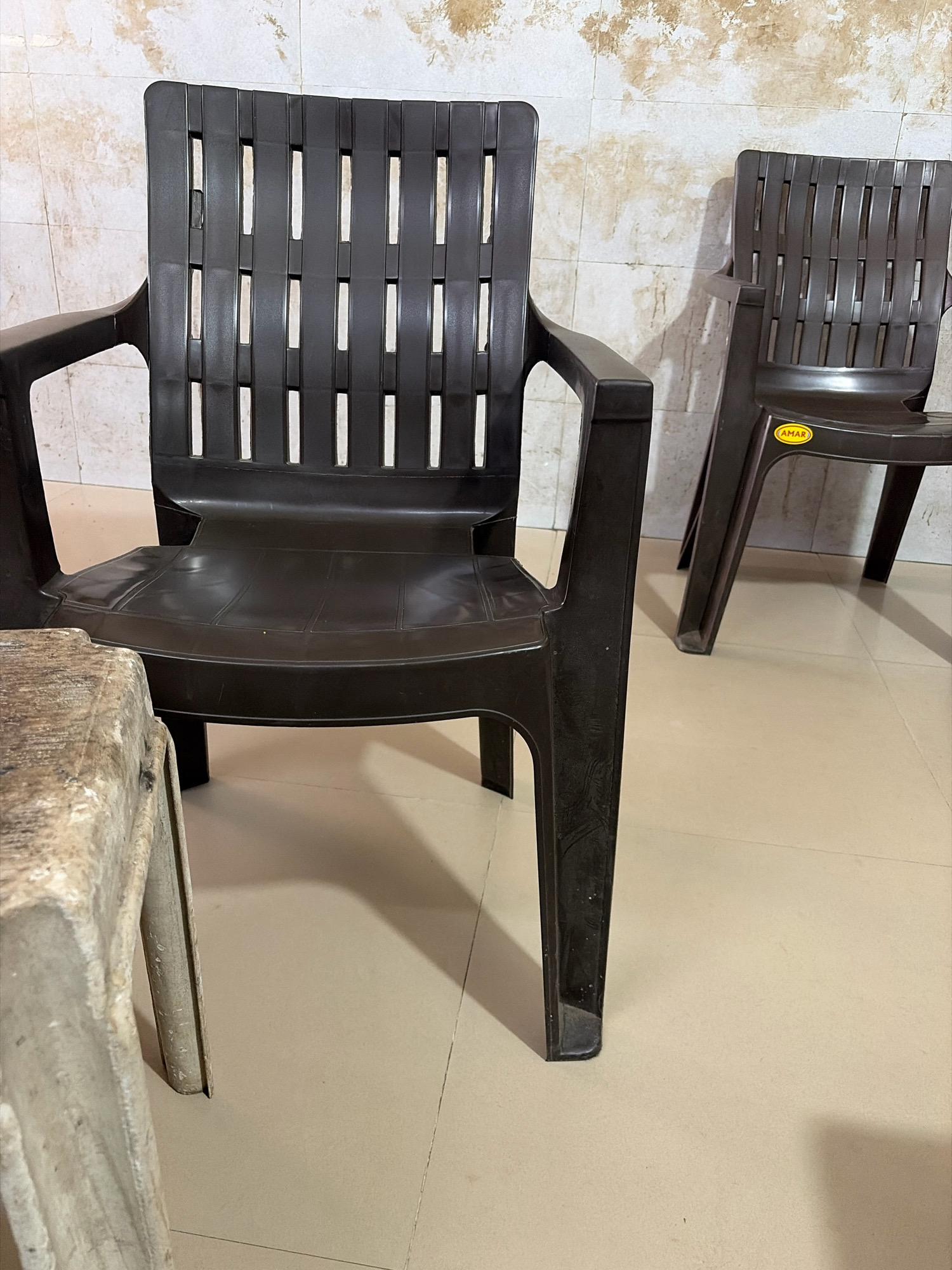 Two dark brown plastic chairs are positioned in front of a tiled wall. The chair in the foreground is on the left side of the image, and the chair in the background is on the right. The chair on the right has a yellow oval sticker with the word "AMAR" printed on it. The floor is tiled in a light beige color. A portion of a light-colored, textured object, possibly a table, is visible in the lower left corner of the image. The scene appears to be an outdoor or semi-outdoor area. The city of Himatnagar, India, is not depicted in this image.