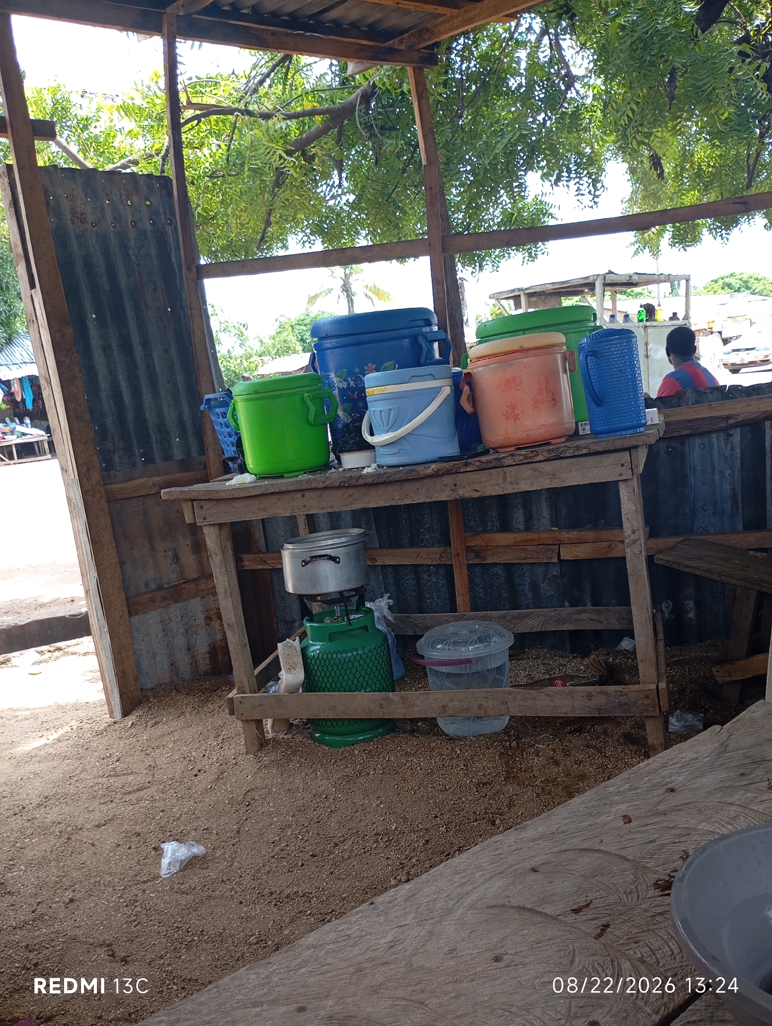 The image depicts an outdoor stall in Shendam, Nigeria. The stall is constructed with corrugated metal and wooden planks, with a rustic wooden table supporting several large plastic containers of various colors, including blue, green, and orange. Some of these containers appear to be coolers or water dispensers. A silver pot sits on top of a green gas cylinder. The ground is covered with sand and wood shavings, with some scattered debris. In the background, trees with lush green foliage are visible, suggesting a tropical or subtropical environment. A person is partially visible in the background, wearing a red and blue striped shirt, facing away from the camera. The overall scene suggests a casual, possibly commercial, setting typical of a local market or roadside vendor.