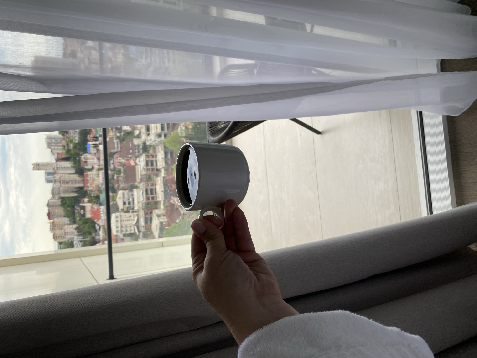 A hand holds a white mug in front of a window, revealing a cityscape and part of a balcony. The cityscape is of Sochi, Russia, characterized by densely packed buildings and lush greenery. The mug contains a dark liquid, presumably coffee or tea. The balcony has a light-colored floor and a railing. Light-colored curtains are partially drawn, obscuring part of the view. The person wearing a white bathrobe.