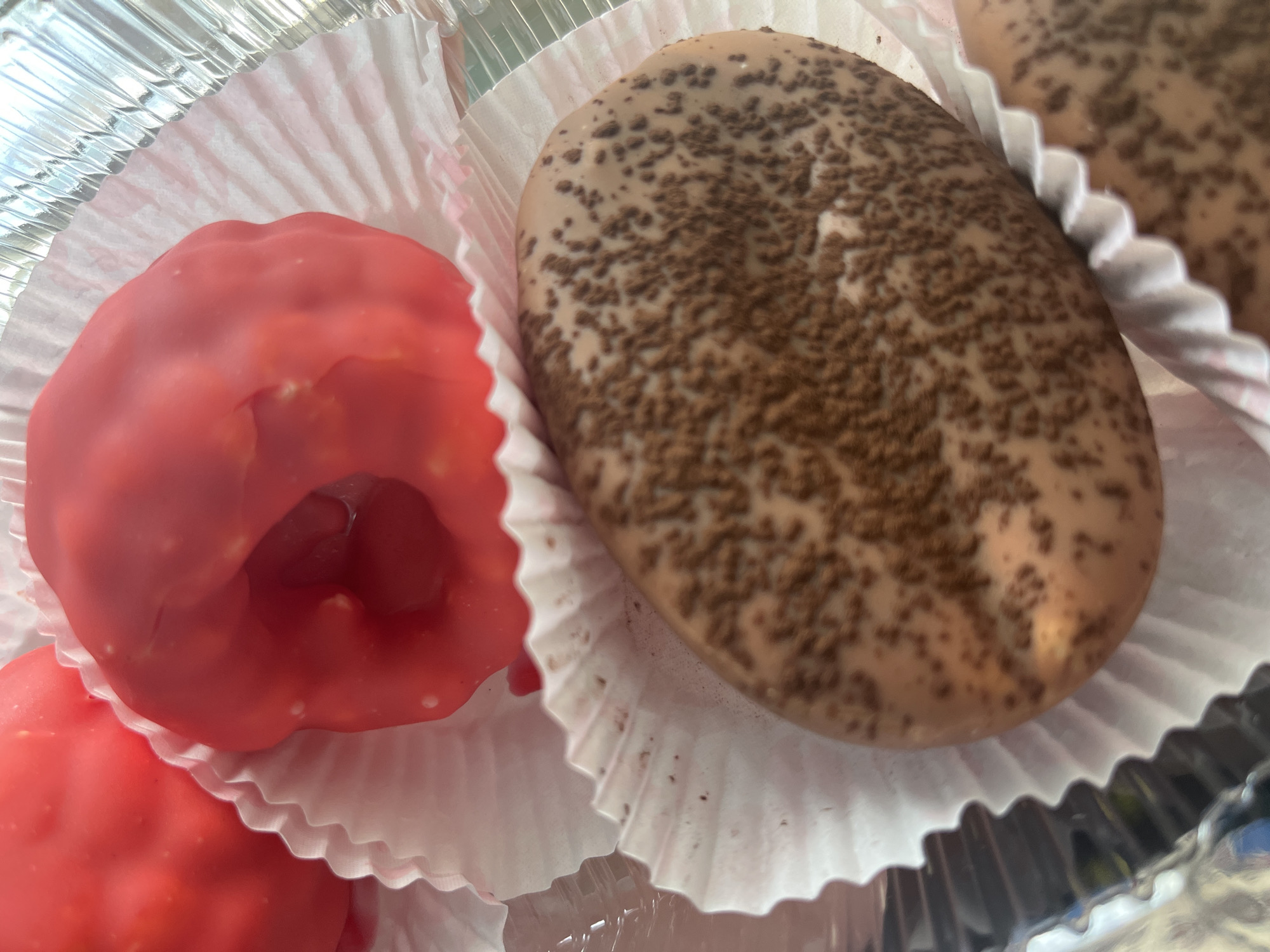 This is a close-up, eye-level shot of two red, round pastries with a hole in the center, sitting in white paper cupcake liners. To the right of the red pastries, there is a large, oval-shaped pastry with a light brown coating sprinkled with dark brown particles. The pastries are arranged in a foil baking pan. The background is out of focus, but appears to show the edge of the pan and possibly other pastries. There is no visible indication of people, actions, or interactions, and no discernible environmental context that would suggest a specific location within the city of Desyatikhatka, Russia.