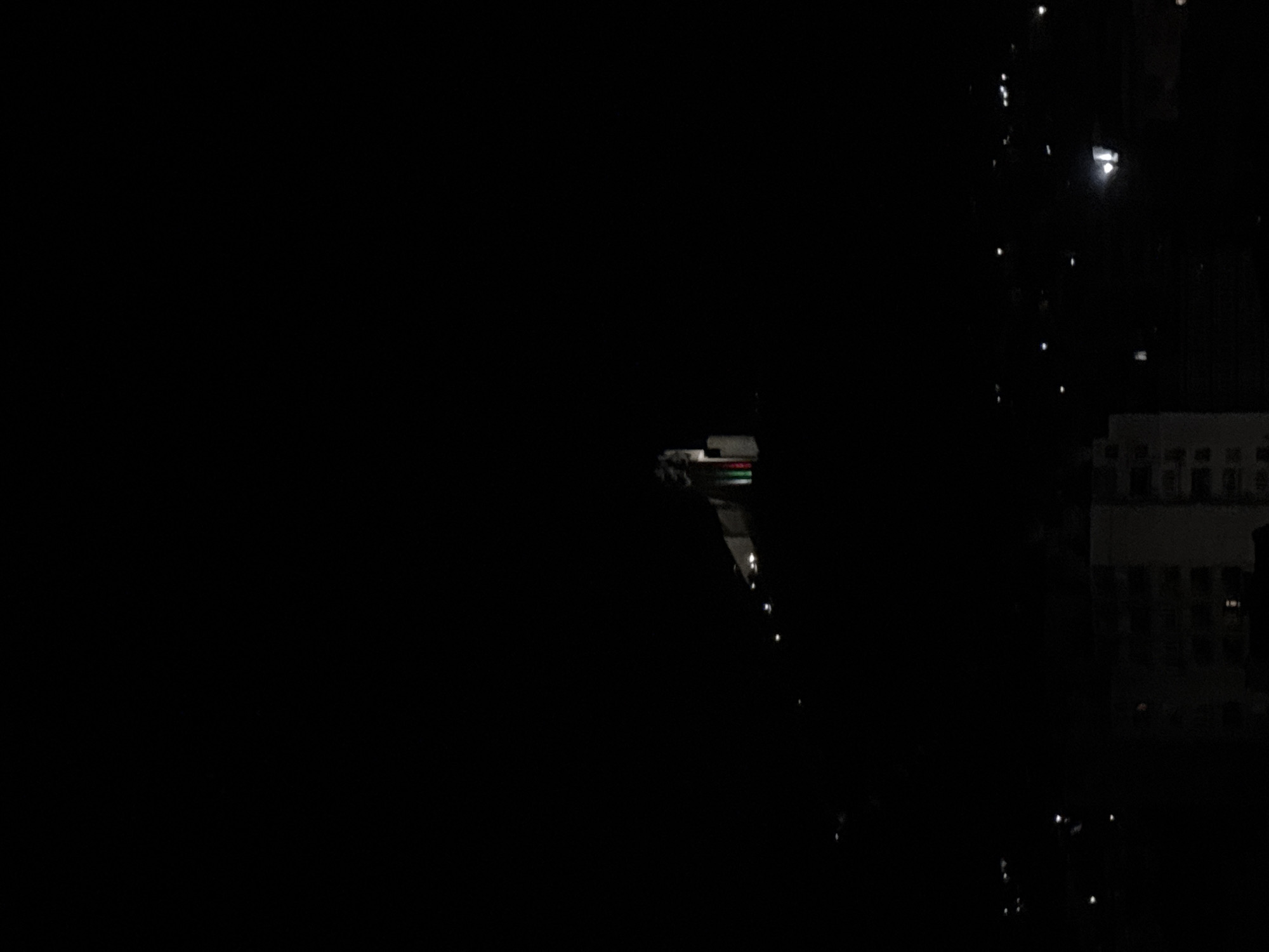 This image shows a dark, nighttime scene. On the right side, a building with illuminated windows is visible. In the middle distance, there is a structure with white and colored lights, possibly a bridge or elevated roadway. The foreground is dark, suggesting a street or area with limited lighting. Raindrops or moisture are present on the window or lens, creating blurred light reflections.