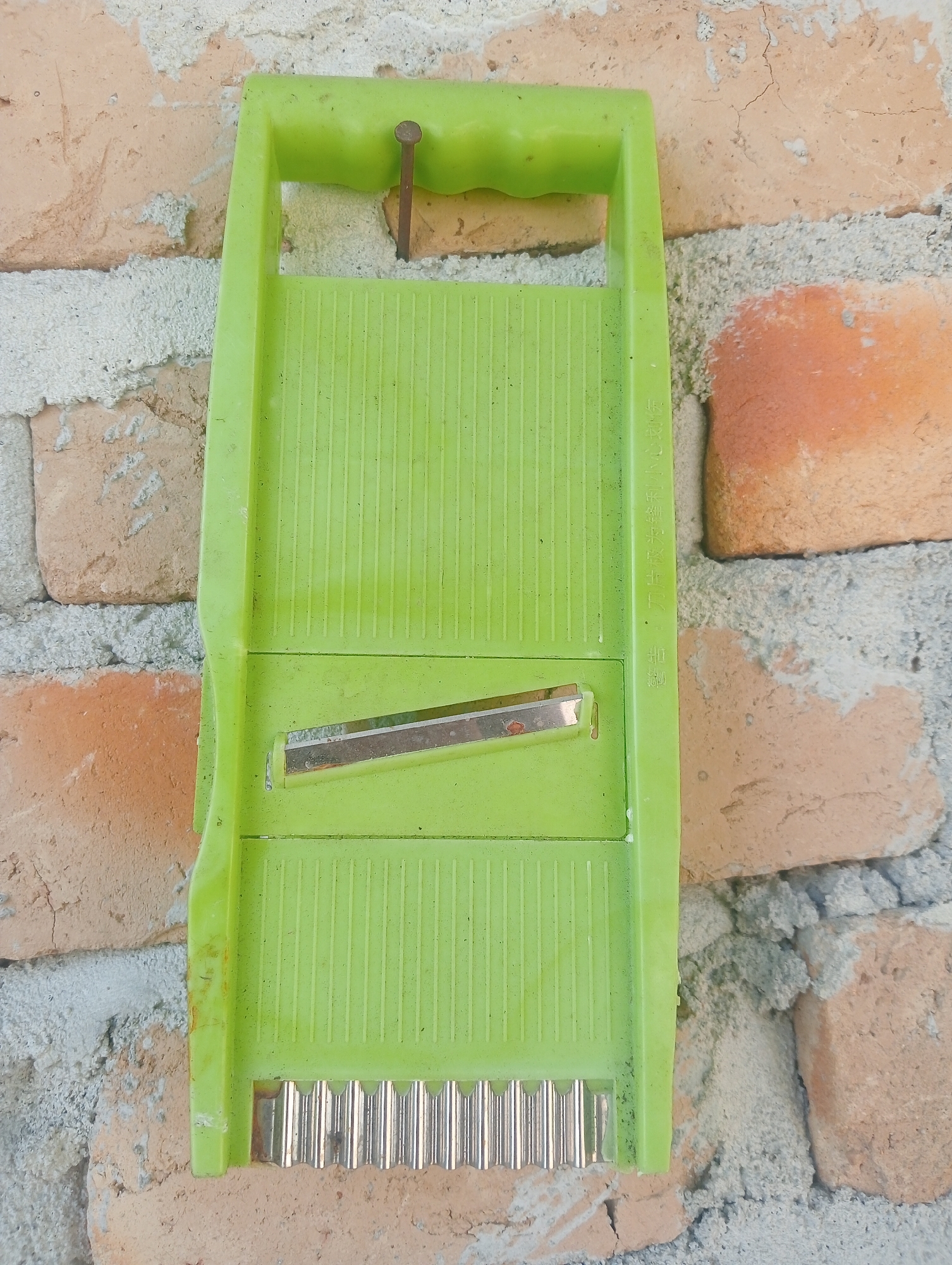 A bright green mandoline slicer is affixed to a brick wall in Kota Negara Timur, Indonesia. The slicer has a metal blade and a textured surface for grating. It is attached to the wall by a single nail driven through its handle. The brick wall is made of rough, uneven bricks in shades of orange and brown, with patches of white mortar. The scene is well-lit, with natural light casting soft shadows.