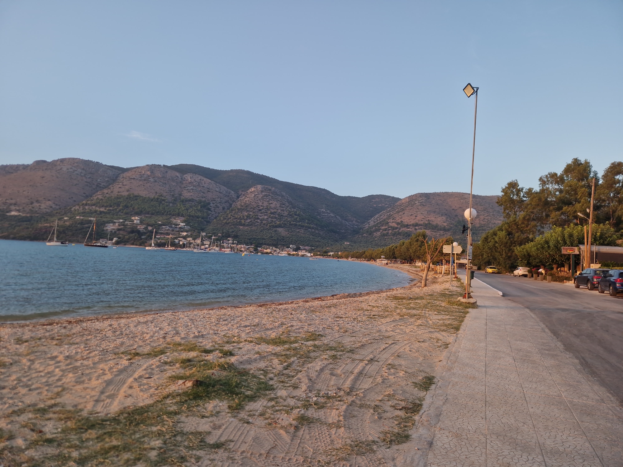 This image depicts a coastal scene in Platamónas, Greece, during what appears to be the late afternoon or early evening. The foreground is dominated by a sandy beach with tire tracks visible in the sand, indicating vehicular passage. Some sparse vegetation is present on the beach. Adjacent to the beach is a paved sidewalk, bordering a road. Several parked cars are visible along the road, including a dark blue car and a dark gray car. Trees line the road and the sidewalk in some areas. A tall lamppost with a square light fixture stands on the sidewalk. In the middle ground, the calm, blue water of the sea stretches out, with numerous sailboats anchored or moored. A town or village is situated along the coastline, nestled at the base of rolling, brown hills. The hills are covered in sparse vegetation and some scattered trees. The sky is clear and a pale blue, suggesting fair weather.