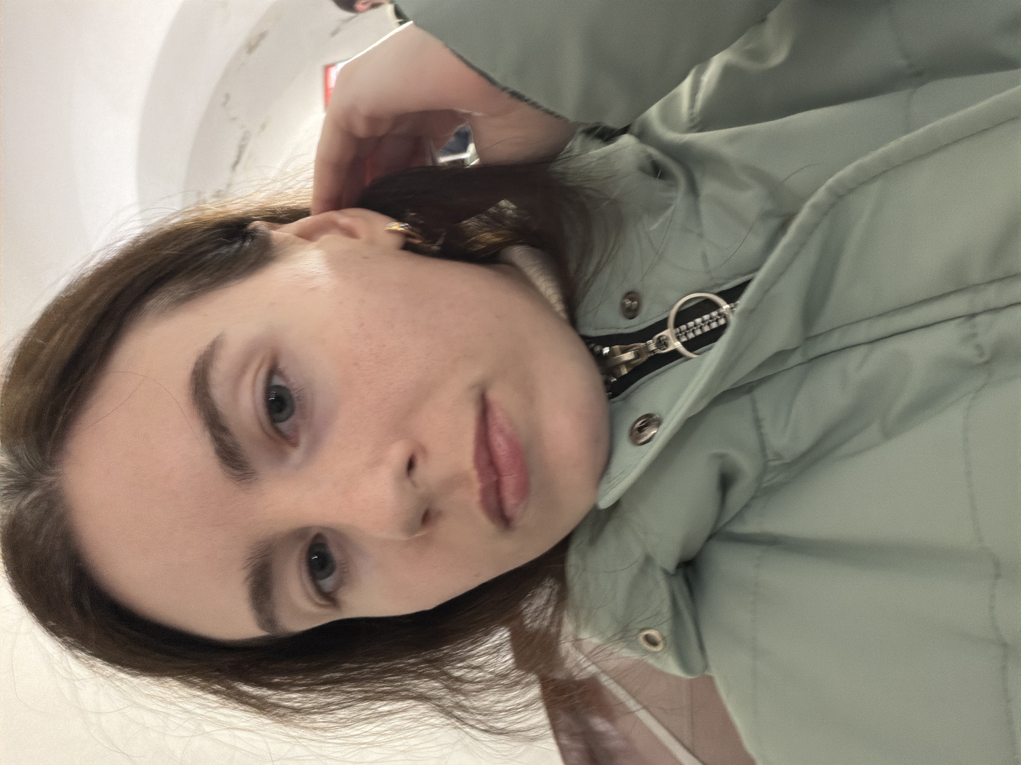 A woman with fair skin, brown hair, and blue eyes is depicted from a close-up, tilted perspective. She wears a light green puffer jacket with a visible zipper and snap buttons. The background is indistinct, with hints of a white, possibly arched structure. Based on details, there is no information regarding the location.