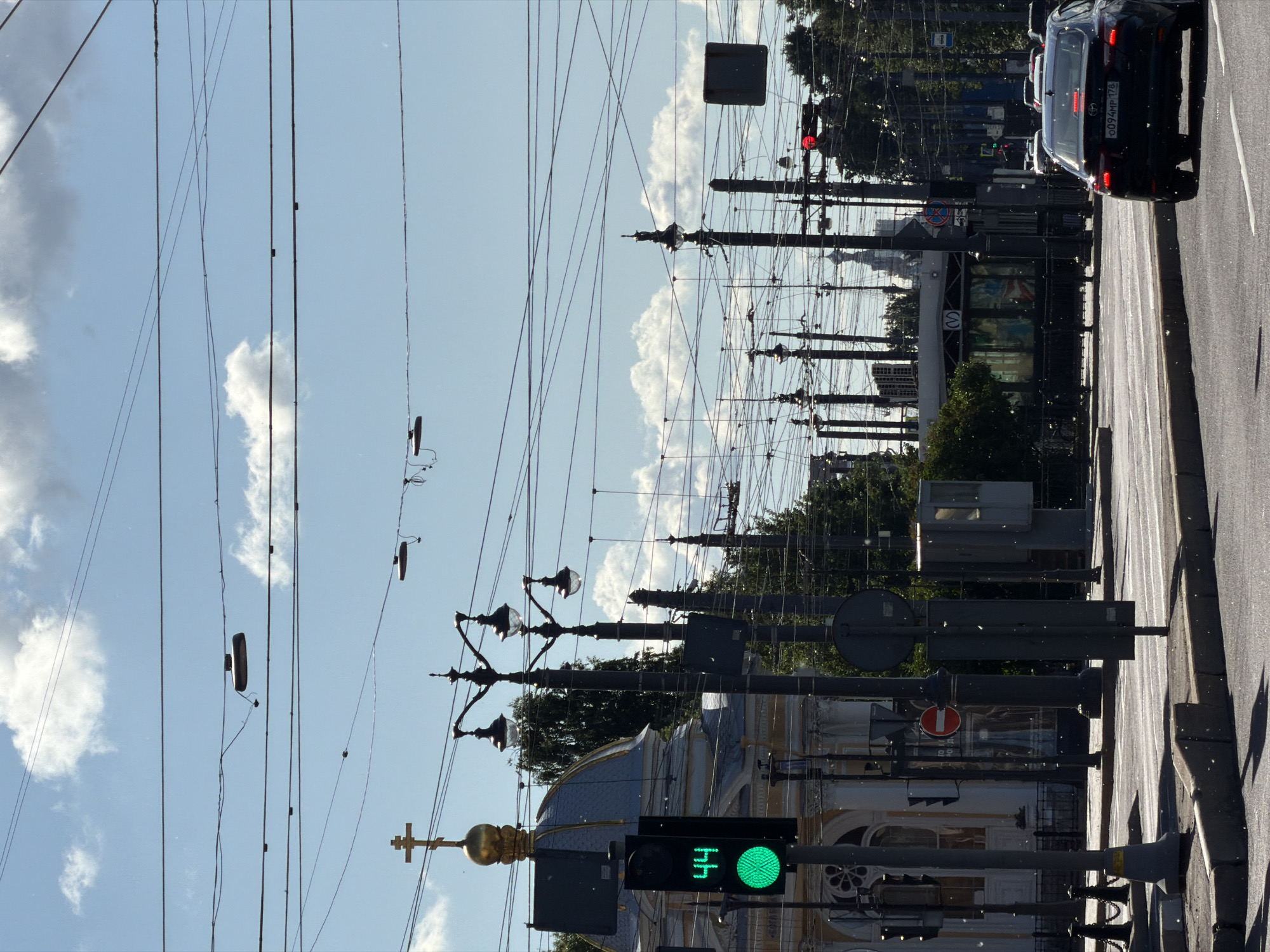 This image depicts a street scene in Saint Petersburg, Russia, on a clear day with some clouds. The perspective is from street level, looking towards the sky and along the street. The most prominent objects are a dense network of overhead wires, likely for public transportation like trams or trolleybuses. Several lampposts with ornate fixtures are visible, as is a traffic light showing a green signal. In the background, trees line the street, and a classical building with a golden dome and cross, characteristic of Russian Orthodox churches, is partially visible. A dark-colored car is in the foreground on the right, and other cars are seen further down the street, suggesting moderate traffic. The lighting suggests it is daytime, likely morning or afternoon, given the bright sunlight and shadows. The overall atmosphere is that of an urban environment. The license plate of the car reads "CO94MP 178".