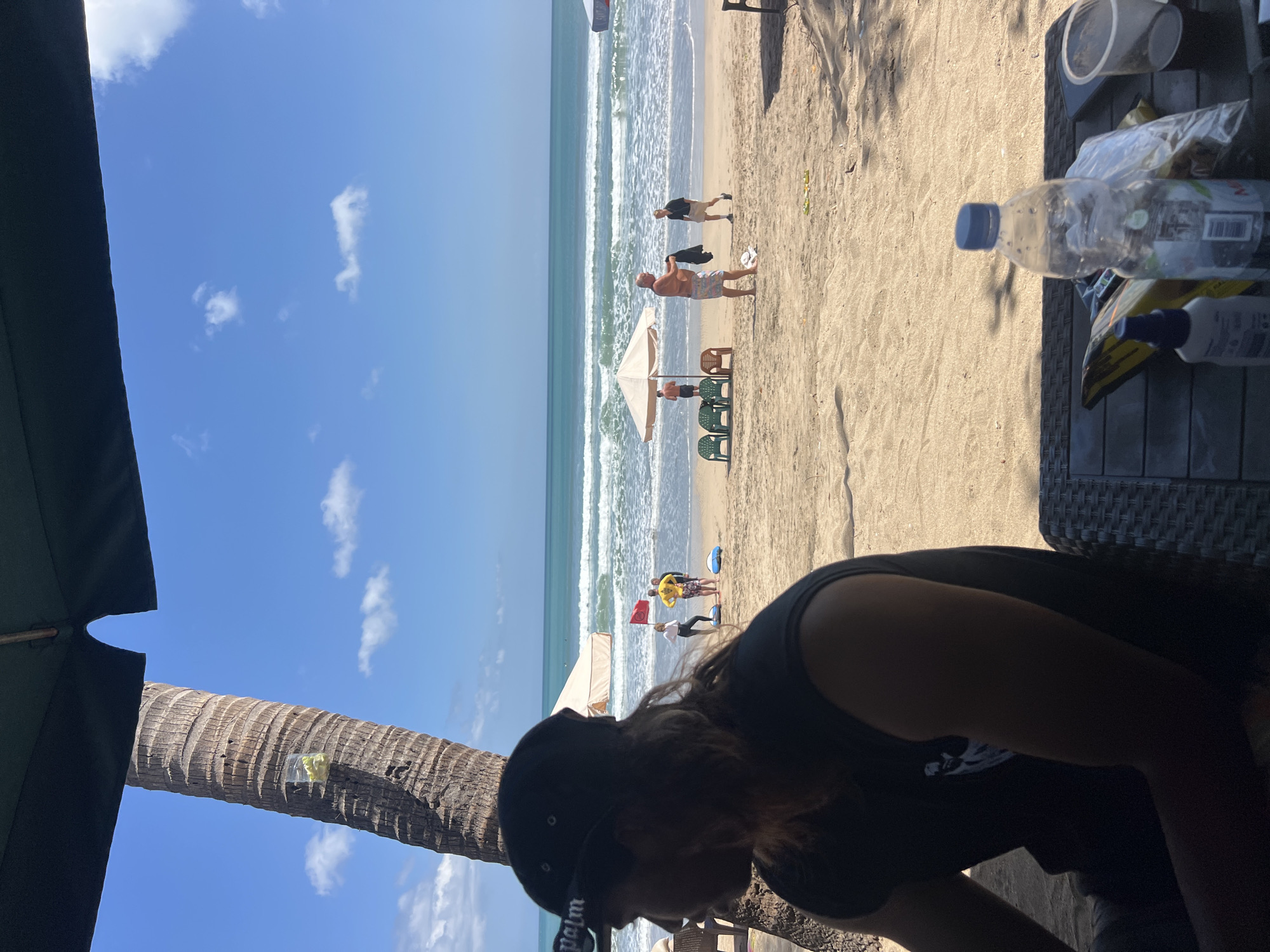 The image depicts a beach scene in Kuta, Indonesia. In the foreground, a person wearing a black sleeveless shirt and a black baseball cap is seated, facing left. Partially visible on the table in front of them are a plastic bottle of water, a spray bottle, a bag of snacks, and a clear plastic cup. A palm tree trunk is prominent on the left side of the frame.
The midground shows a sandy beach with several people. Two individuals are standing near a large tent or umbrella, and a few other people are scattered on the sand, some near beach chairs and umbrellas. Further back, the ocean is visible with moderate waves. The sky is clear blue with scattered white clouds. In the distance, the horizon line separates the ocean and sky. The overall impression is a sunny day at a tropical beach.