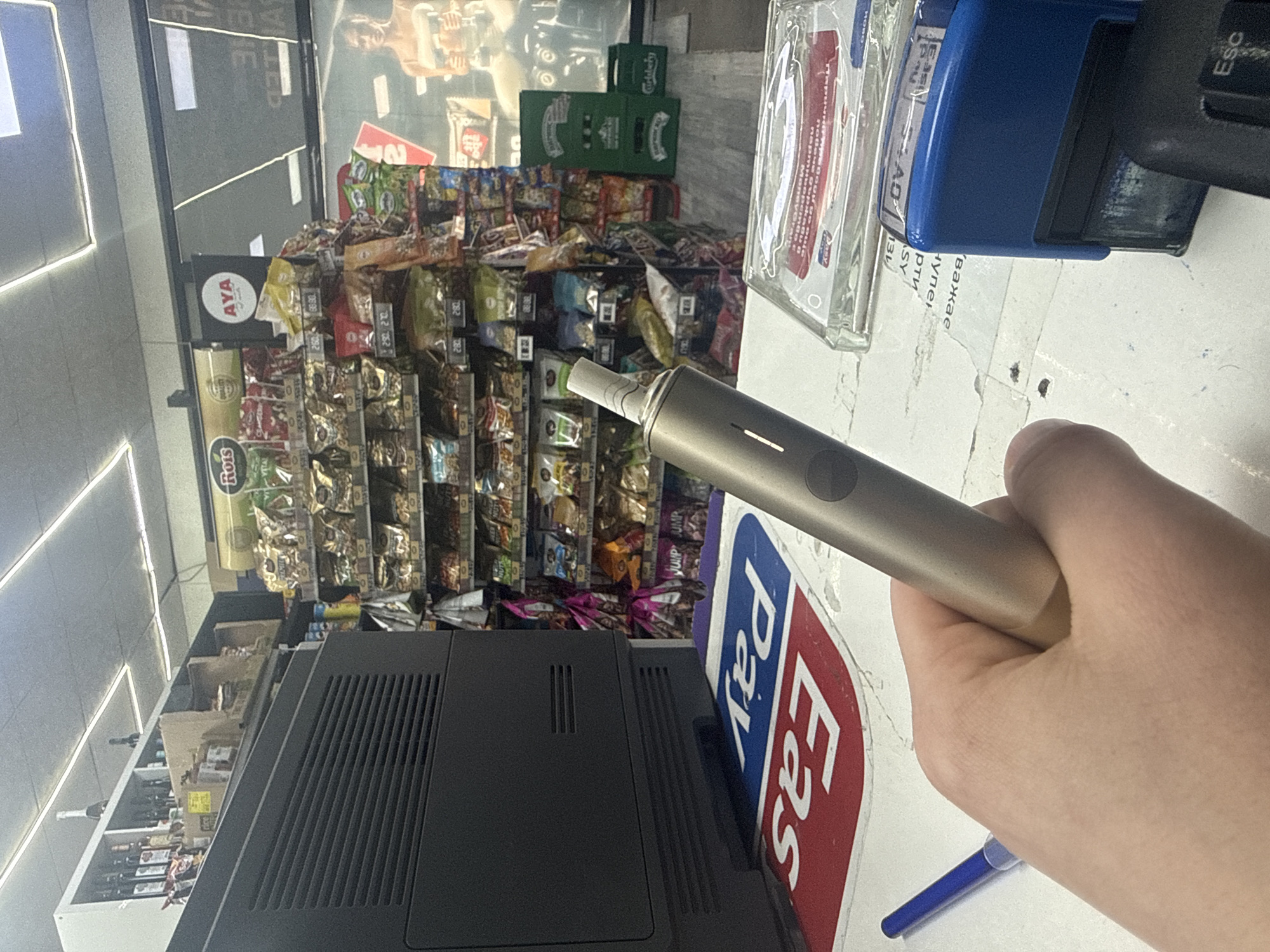 The image depicts a scene inside a store, possibly a convenience store or small supermarket. A person's hand holds a gray electronic device, possibly a vape pen. An "Easy Pay" sign is visible in the foreground. Behind the person's hand are shelves filled with various snacks and food products. Several boxes of Carlsberg beer are also present. Based on the "AYA" sign, the location is likely in Sofia, Bulgaria, as this is a known Bulgarian retail chain. The image likely depicts the point of sale area of a store, near the cash register and checkout counter.