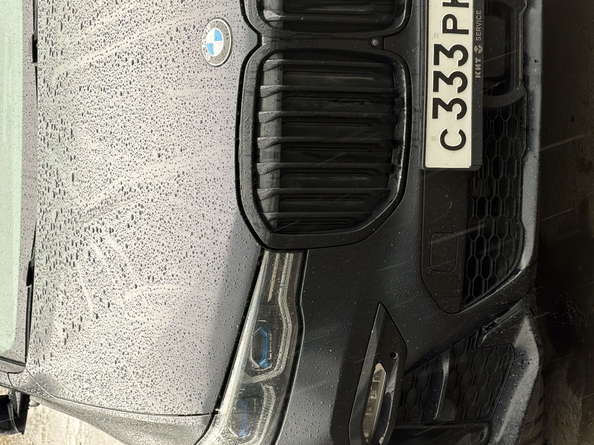 The image depicts the front of a black BMW car, possibly an SUV, with the license plate "C333PH". The car is covered in raindrops, indicating recent or ongoing rain. The BMW logo is visible on the hood above the grill. The grill is a blacked-out vertical slat design. The car is in Izhevsk, Russia, based on the Cyrillic text "КИТ SERVICE" on the license plate. The license plate appears to be a standard Russian license plate. The headlight is partially visible on the left. The bumper has a black honeycomb pattern on either side. The car appears to be parked, but the immediate surroundings are not clearly visible.