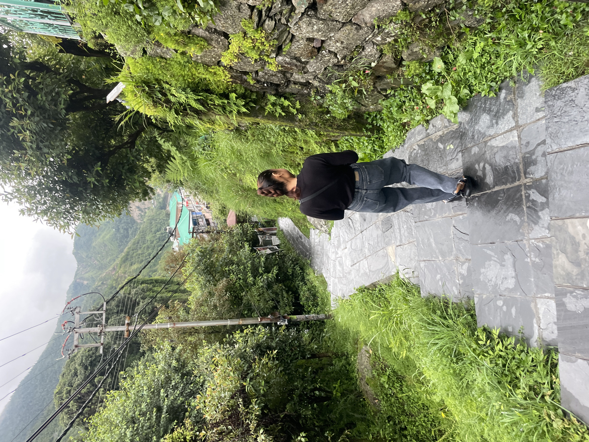 A woman with her hair in a braid walks down a stone staircase in Bhagsu, India. She is wearing a black long-sleeved shirt and grey jeans. The staircase is flanked by lush green vegetation on the right, with a stone wall and a rock face covered in plants. On the left, there is more greenery, a telephone pole with wires, and buildings nestled in the valley below. In the background, misty mountains rise. The scene suggests a slightly overcast day with damp stone steps, indicating recent rain.