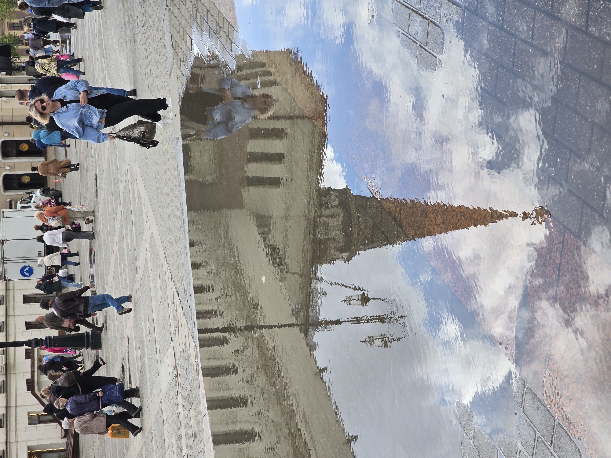 The image captures a street scene in Moscow, Russia. A puddle on the cobblestone pavement reflects the sky, clouds, and a building with a prominent spire, possibly a part of the Kremlin. A group of people is walking along the pavement near the puddle, seemingly oblivious to the reflection. The people are dressed in casual clothing, and the scene suggests a normal day in the city with pedestrians moving about their daily activities.