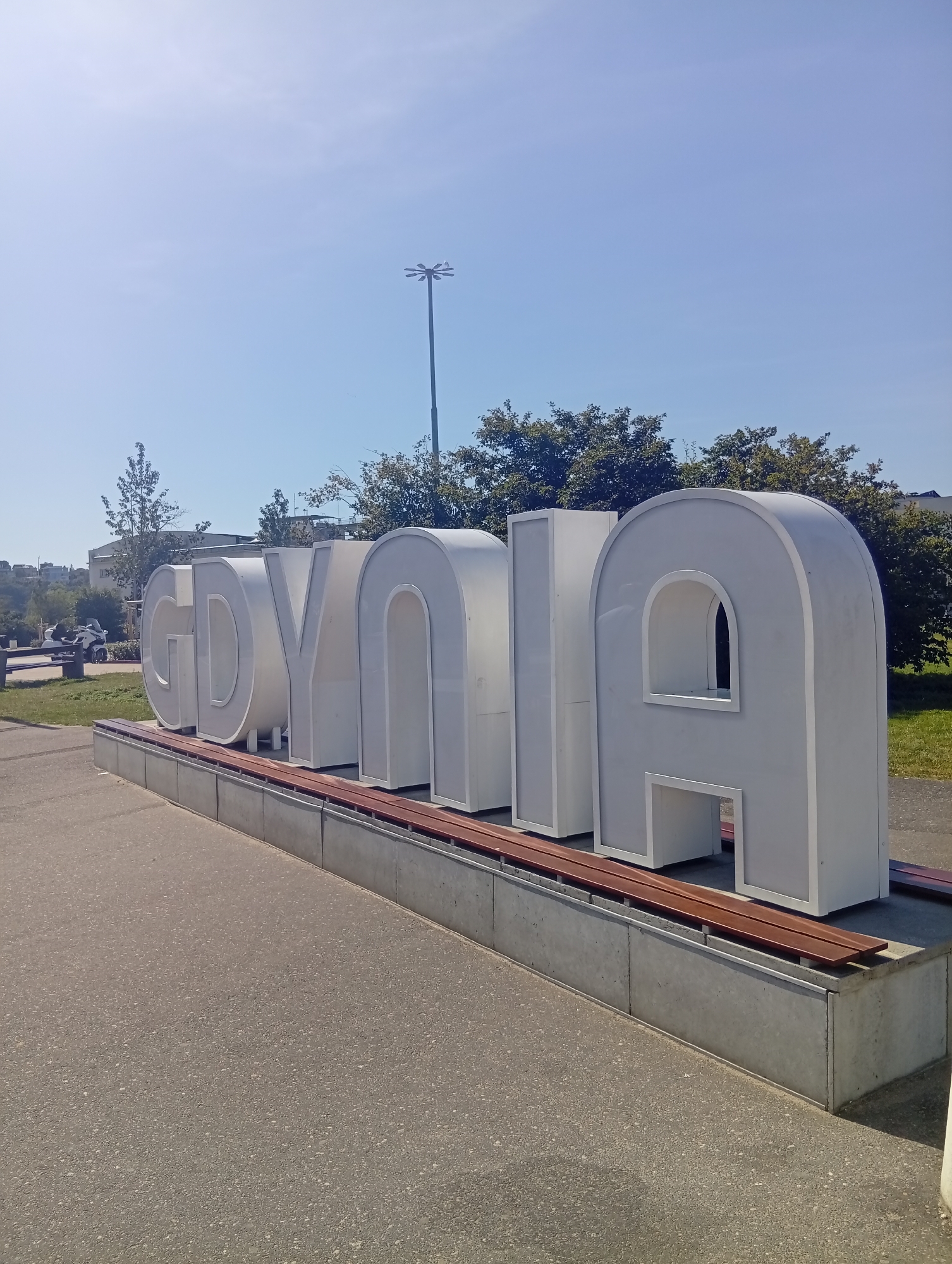 The image shows a large, white, three-dimensional sculpture spelling out "GDYNIA" in front of a clear blue sky on a sunny day. The letters are spaced apart and stand on a concrete base with wooden benches. Behind the sculpture, there are trees and some buildings. The ground in front of the sculpture is paved. The sculpture is located in the city of Gdynia, Poland. There are no people or animals visible in the image. The scene appears to be a public outdoor space, possibly a promenade or park area. The lighting indicates it is daytime.