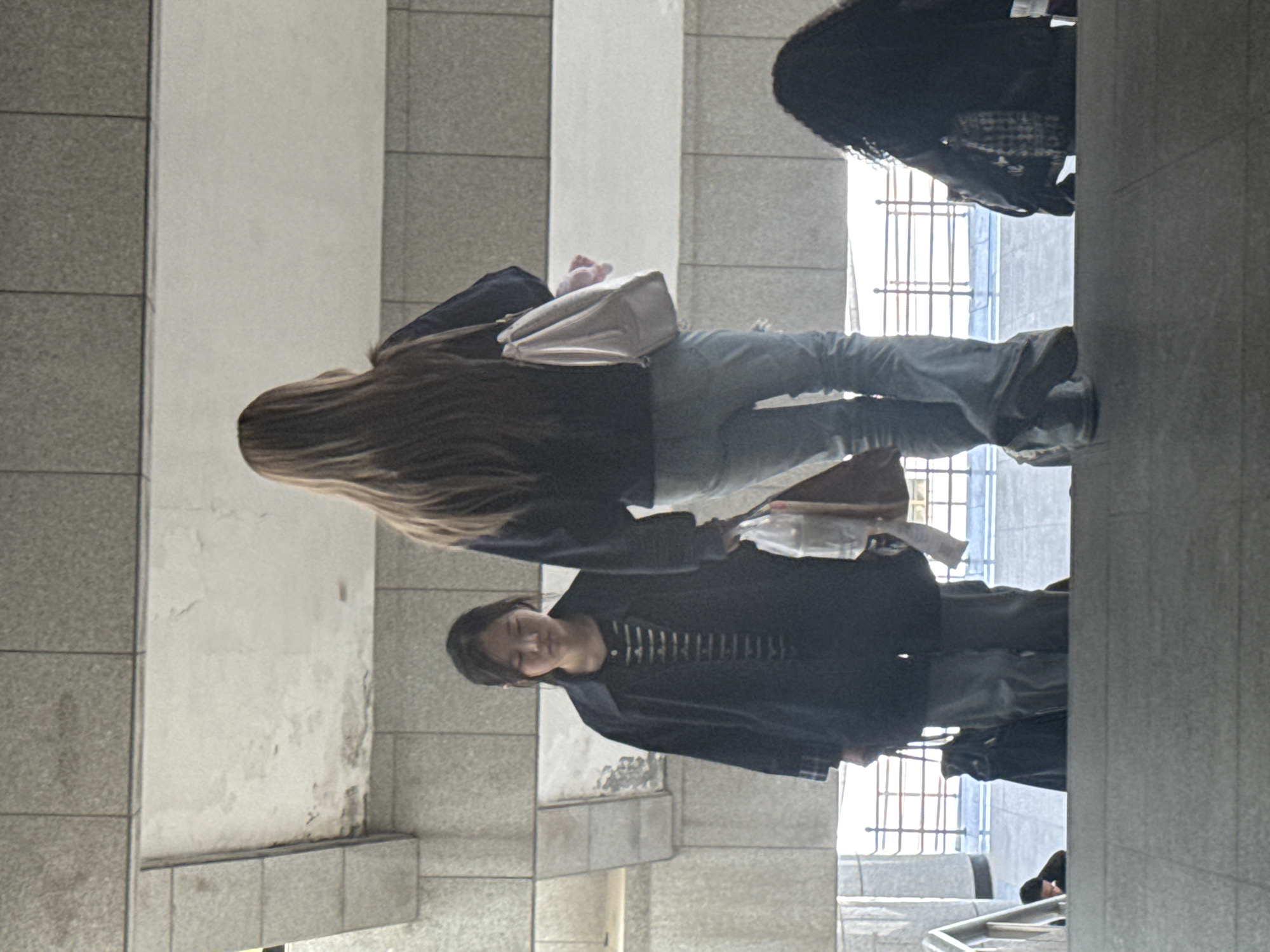 The image shows an indoor scene, potentially a subway station or a covered public walkway in Zhongtun, China. Three individuals are present. The person closest to the camera, a woman, is wearing a dark jacket over a striped shirt and light-colored jeans. Another person, a woman with long hair, is wearing a dark jacket, light-colored jeans and carrying a light-colored bag. A third person, also a woman with dark, curly hair, is wearing a backpack. The background includes tiled walls and what appear to be metal bars, suggesting a station or public area. The lighting is relatively dim.