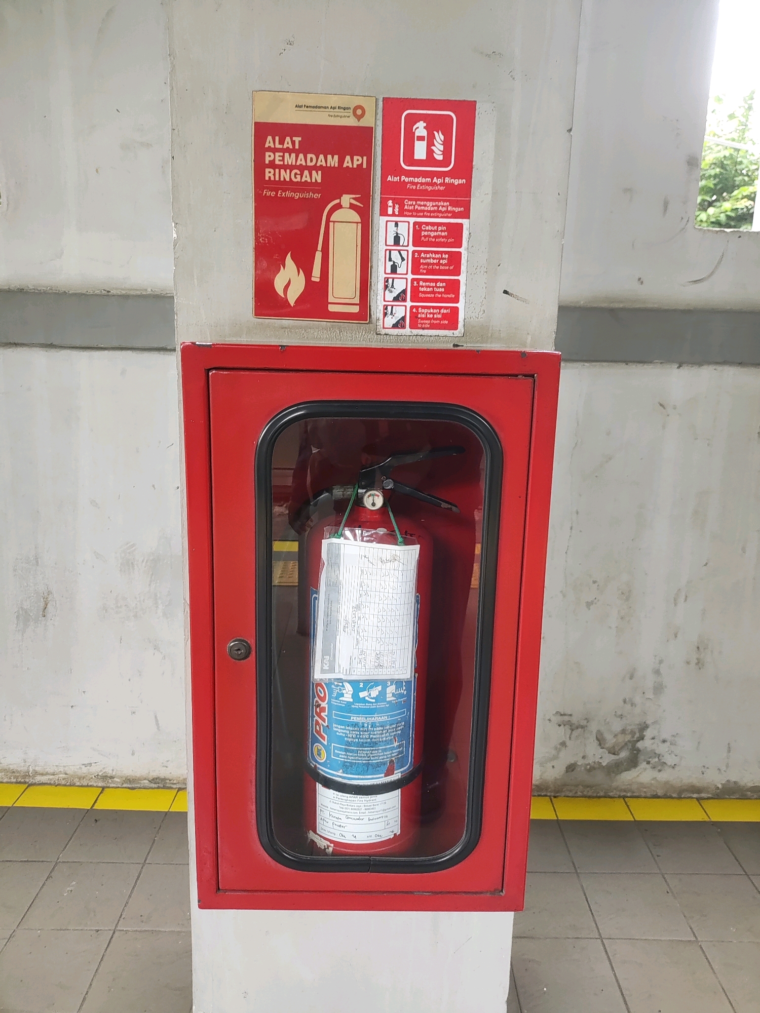 In Telagamurni, Indonesia, a red fire extinguisher cabinet is mounted on a concrete pillar. The cabinet has a glass door, and inside, a fire extinguisher is visible. Two red and yellow warning signs are affixed to the pillar above the cabinet. The signs contain text in Indonesian and English, with the left sign showing a graphic of a fire extinguisher and flames, and the right sign illustrating the steps for using a fire extinguisher. The environment appears to be an indoor or semi-enclosed public space with tiled flooring and a yellow striped boundary line along the bottom.