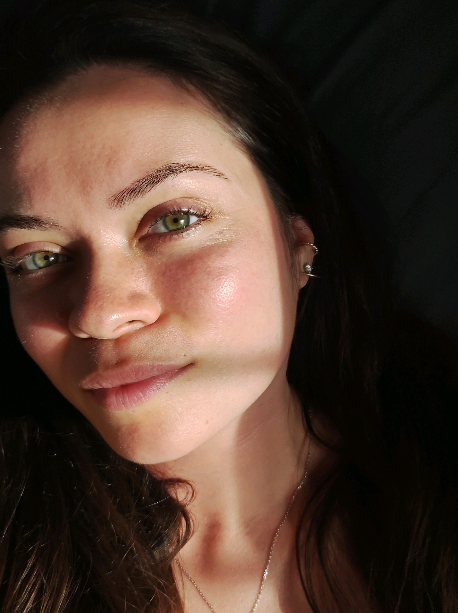 A close-up, head-and-shoulders portrait of a young woman with medium-toned skin and long, dark brown hair. Her eyes are a striking green, and her eyebrows are well-defined. Her lips are slightly parted. She is wearing a thin, silver chain necklace. The lighting creates a dramatic effect, with a strong shadow cast across her forehead and cheek. A thin, metallic earring is visible on her right ear. The background is dark and out of focus, suggesting an indoor setting. There are no discernible landmarks or contextual clues to indicate a specific location within Chaikovsky, Russia.