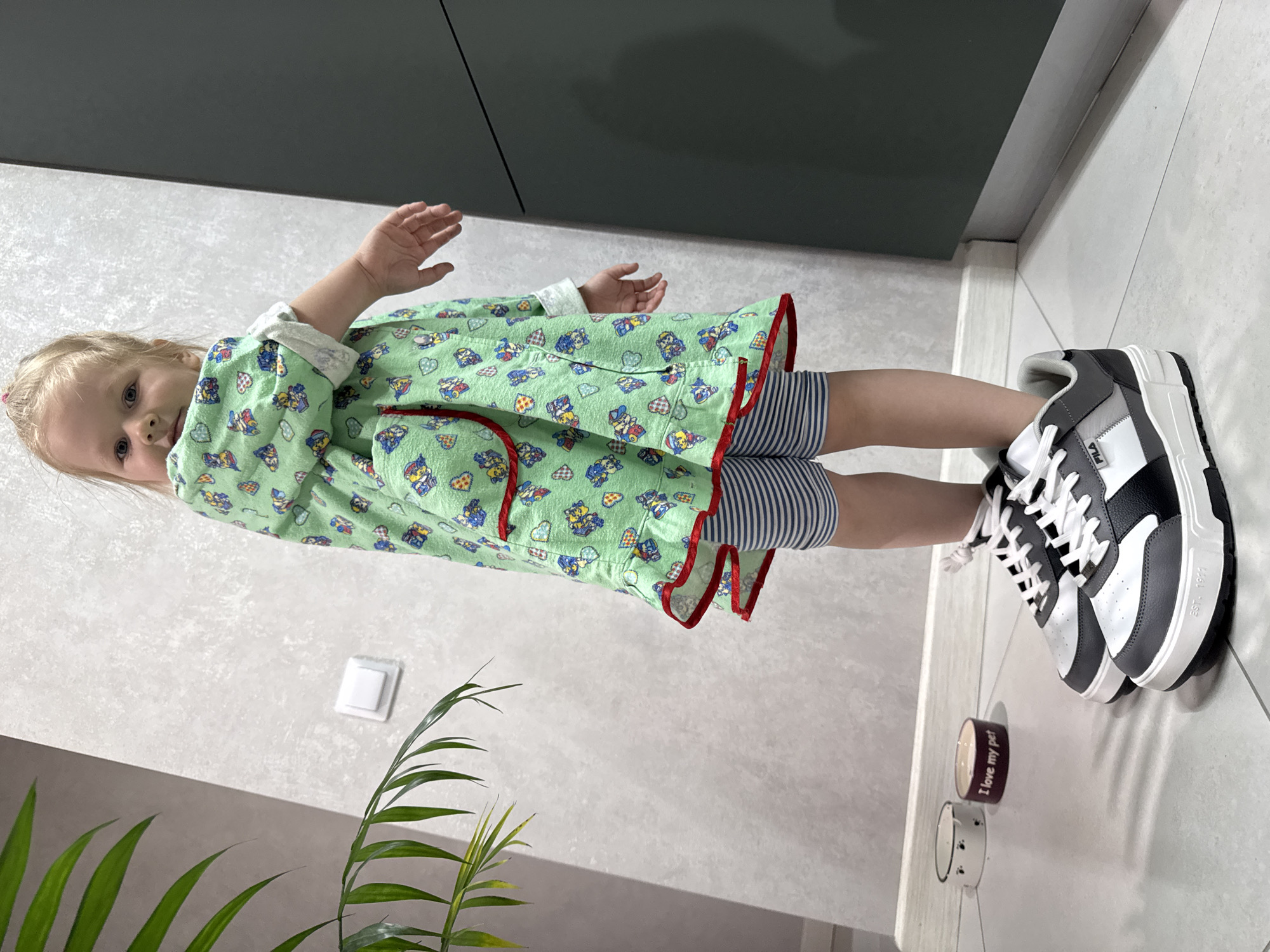 A toddler stands in what appears to be an interior space, possibly a hallway or room within a building in the Dvurechenskoe S/P area of Gornyi, Russia. The child is wearing a green robe with cartoon-like patterns and striped shorts. They are also wearing large, black and white athletic shoes. The child's arms are partially raised. Near the shoes are two pet bowls. The wall behind the child is a light grey and there is a plant with long green leaves in the foreground. A dark grey cabinet or wall panel is visible in the background. A light switch is mounted on the grey wall. The floor is covered with white tiles.