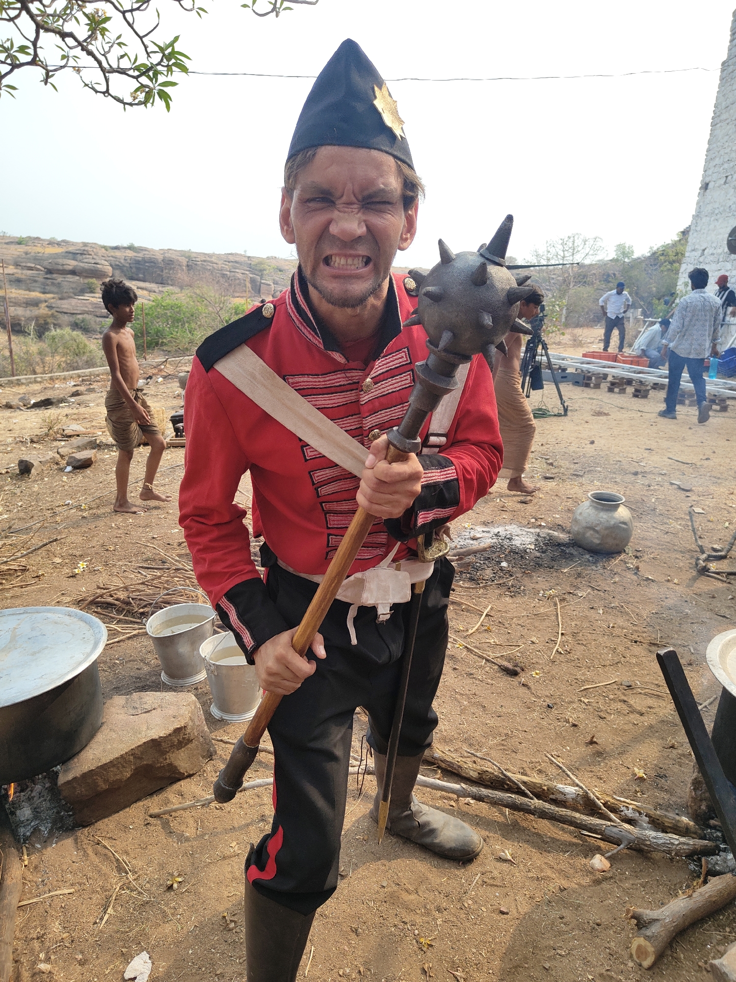 The image depicts a man dressed in a historical British soldier's uniform, including a red jacket, black pants, boots, and a black hat. He is holding a spiked mace and making a face with clenched teeth. The location appears to be an outdoor setting in Dupadu, India, with a landscape of rocky hills and sparse vegetation visible in the background. Several people are present in the background, including a shirtless boy and individuals who appear to be part of a film crew. Various objects, such as cooking pots, buckets, firewood, and a camera tripod, are scattered around the scene. Smoke from a fire is also visible. The setting suggests a film shoot or historical recreation event.