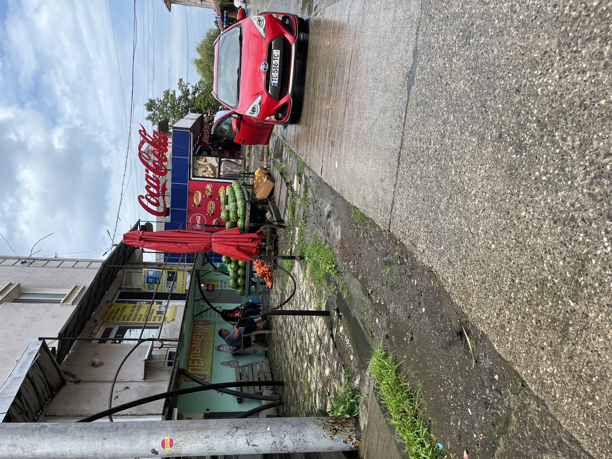 The image shows a street scene in Poti, Georgia, on a cloudy day. A red Toyota Aqua is parked with its driver's door open on the right side of a wet, concrete road. A small market stall selling watermelons is set up to the left, with a red umbrella providing shade. Behind the stall, a building houses several shops, including one with a prominent "Coca-Cola" sign. Two people are seated on chairs outside one of the shops, and another person is visible inside a shop with an open door. The ground is wet, indicating recent rain or damp conditions. Signs in Georgian and Russian are visible on the buildings.