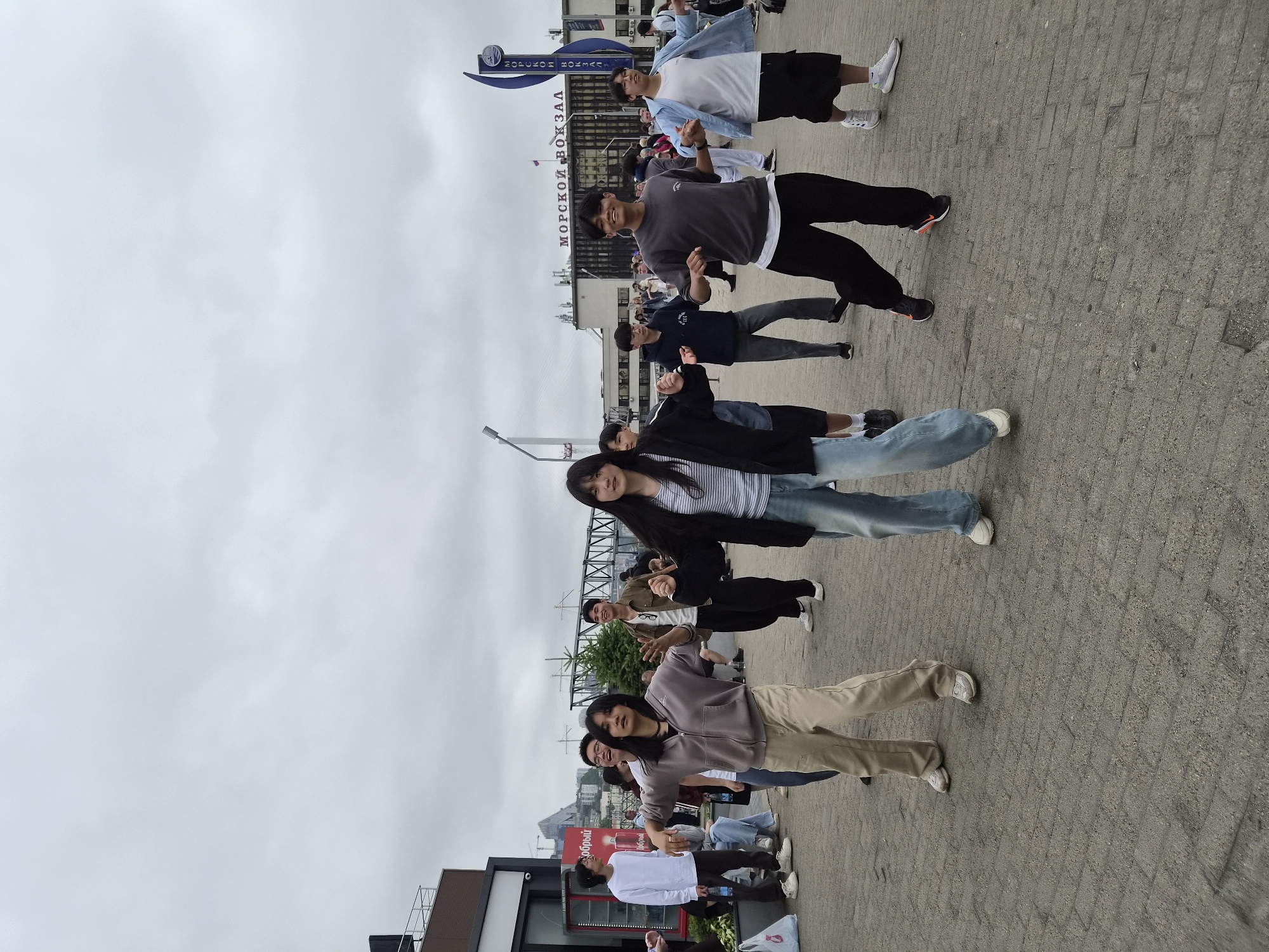 The image captures a group of young people, predominantly of Asian descent, engaged in a dance performance on a paved outdoor plaza. The scene is set against the backdrop of a cloudy sky and urban architecture, specifically identified as the Vladivostok Sea Passenger Terminal (МОРСКОЙ ВОКЗАЛ) in Vladivostok, Russia. The building features large windows and signage in Russian.
In the foreground, several individuals are actively dancing, with their bodies in various dynamic poses, suggesting choreographed movement. The group consists of both males and females, dressed in casual, contemporary clothing such as hoodies, t-shirts, jeans, and sneakers. One female in the center wears a grey zip-up hoodie and tan pants, while another on the far right has a black jacket over a striped shirt and jeans. A male in the center-right wears a grey t-shirt and black pants, and another on the far right wears a light blue jacket over a white t-shirt and black shorts.
In the middle ground, more individuals are either participating in the dance or observing. The environment is an open, paved area with textured cobblestone-like surfacing. To the left, a red vending machine and a portion of a modern building with a glass facade are visible. Further in the background, a large bridge structure, power lines, and hills with buildings can be seen, typical of a port city. The overall atmosphere suggests a public event or spontaneous performance in a bustling urban setting.