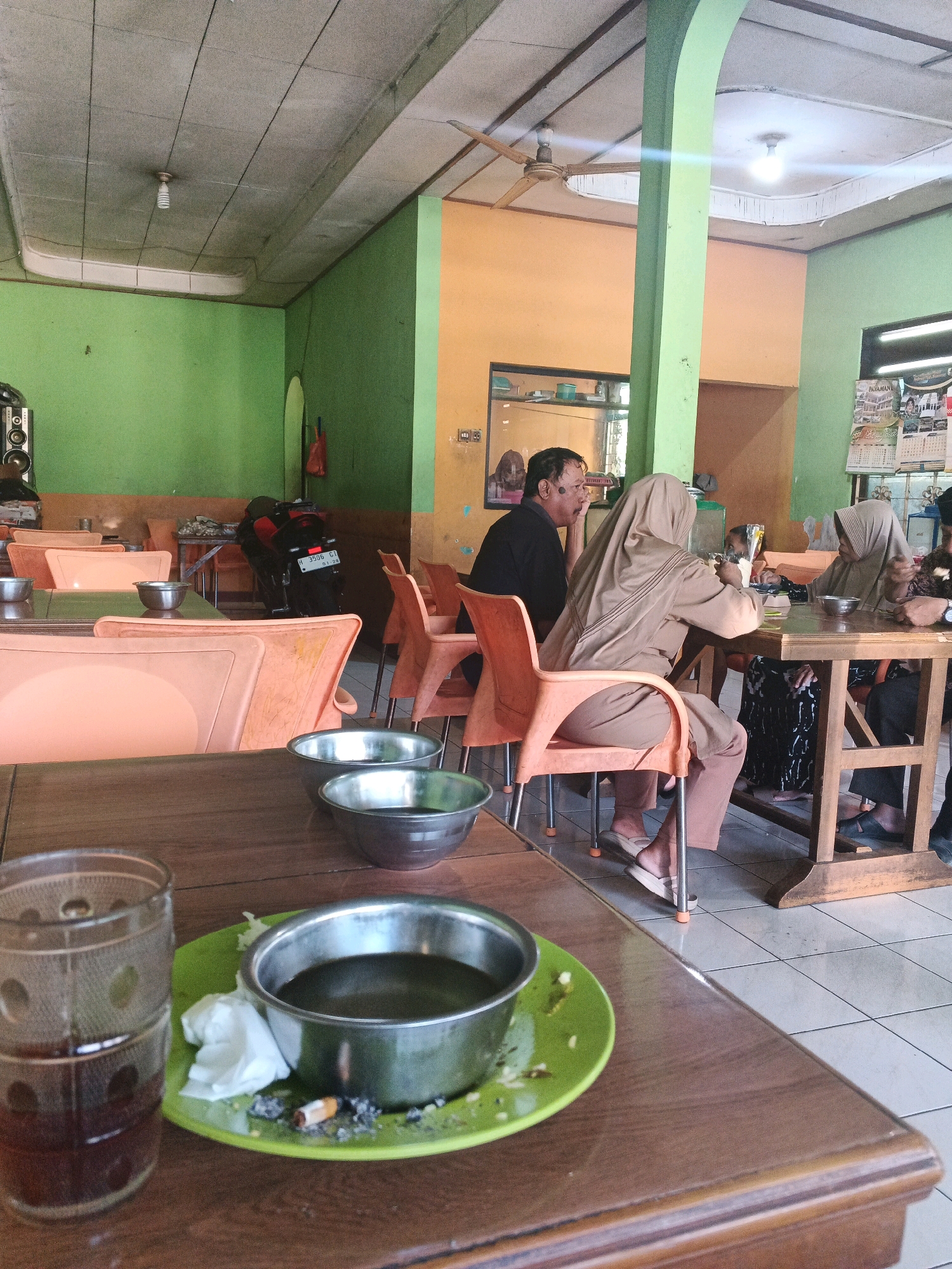 The image depicts the interior of a restaurant in Ungaran, Indonesia, likely a local eatery. The walls are painted in bright green and orange, with a simple, functional ceiling. Several tables are visible, some occupied by patrons. In the foreground, a wooden table holds a glass of dark liquid, a green plate with a bowl of food, a crumpled tissue, and cigarette butts. Further back, a group of people are seated at a table. A man in a black shirt is talking to a woman wearing a light brown hijab, who is also holding a utensil. Another woman in a similar hijab is seated opposite them. The overall atmosphere appears casual and lived-in, with evidence of food and drink service. A motorcycle is parked in the background, suggesting it's a common mode of transport for customers.