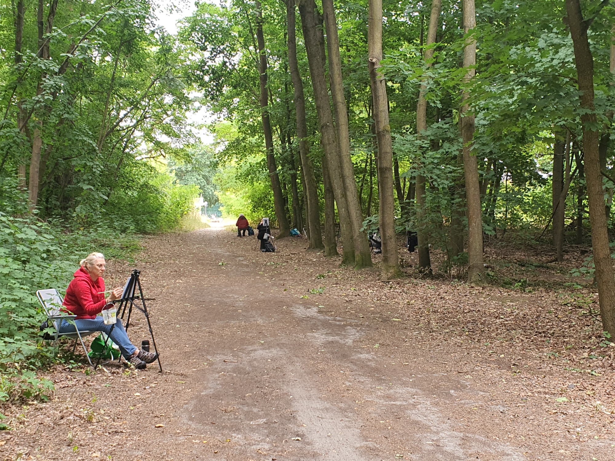 In a woodland park in Falkensee, Germany, a group of people are engaged in an outdoor painting session. The scene is dominated by a dirt path winding through a dense forest with tall, mature trees. In the foreground, on the left side of the path, a woman with blonde hair tied back, wearing a red hooded jacket and blue jeans, is seated in a folding chair. She is actively painting on an easel set up in front of her, holding a paintbrush and a palette. Next to her chair, a green bag rests on the ground.
Further down the path, several other individuals are visible, also positioned along the edges of the path and seemingly involved in painting or observing. One person in a red jacket is near the center, and other figures in darker clothing are scattered amongst the trees. The ground is covered with fallen leaves and dirt, typical of a forest floor. The overall atmosphere is one of quiet, creative activity amidst nature.