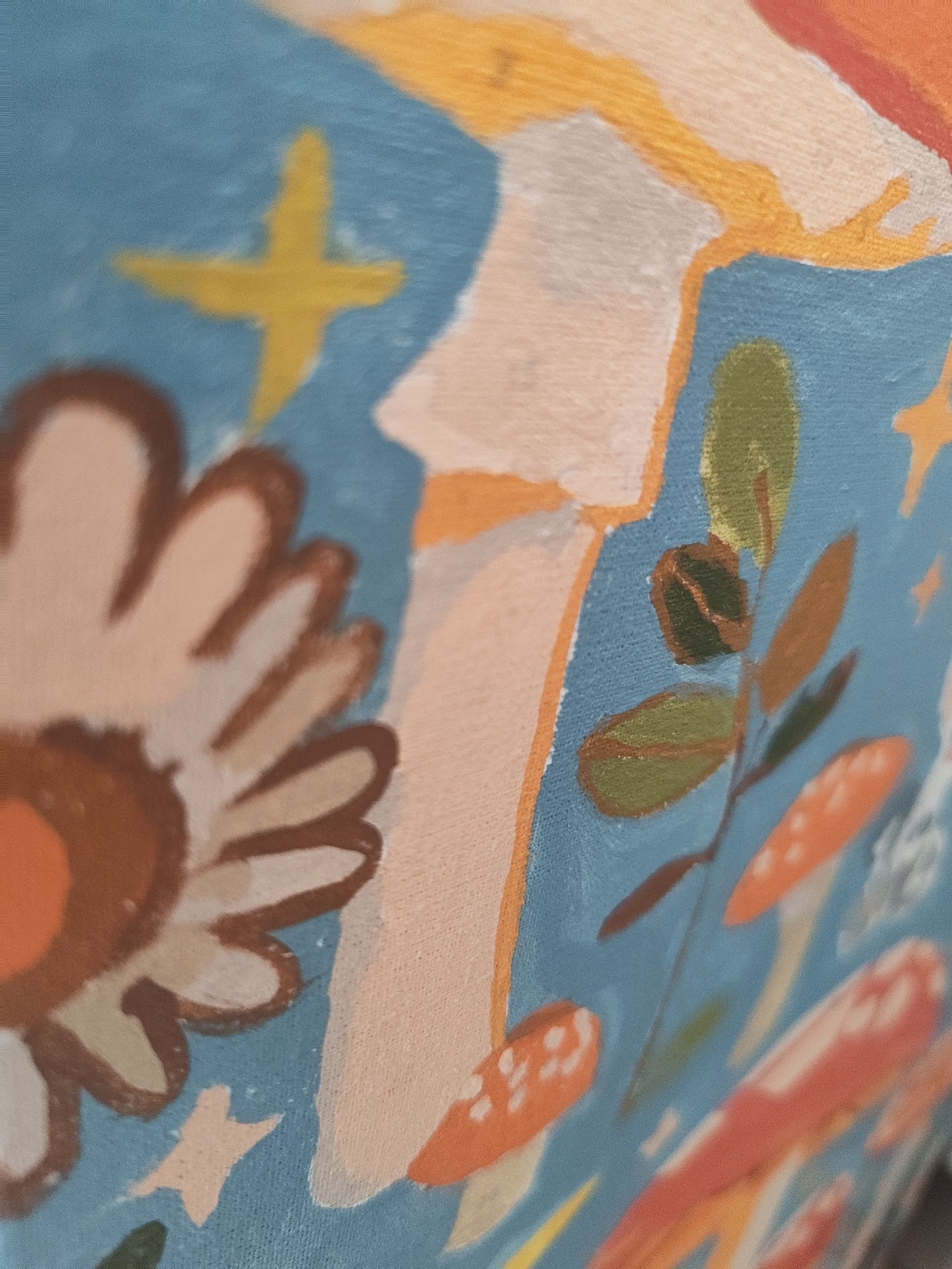 The image is a close-up, shallow depth of field shot of a painting. The painting features a stylized, abstract design with a blue background. Visible elements include a large, brown and cream-colored flower on the left, a yellow star shape above and to the left of the flower, and a cluster of green leaves on a branch with brown outlines to the right of the flower. Several orange and white spotted mushrooms are also depicted. The painting appears to be rendered in acrylics on canvas, with visible brushstrokes and texture. There is no information in the image that provides context about its location in Il’inskiy, Russia.