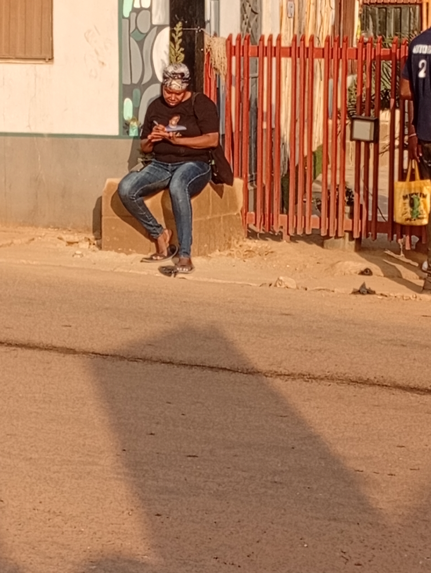 A woman sits on a concrete block beside a road in Narayi, Nigeria. She is wearing a black t-shirt, blue jeans, and sandals, and has a patterned headscarf. She is looking down at something in her hands, possibly a phone. Behind her is a building with a window and a decorative painted wall. To the right is a red metal fence, and part of a person with a blue shirt and a yellow bag. The road is visible in the foreground, with a large shadow stretching across it. The scene appears to be bathed in warm sunlight.