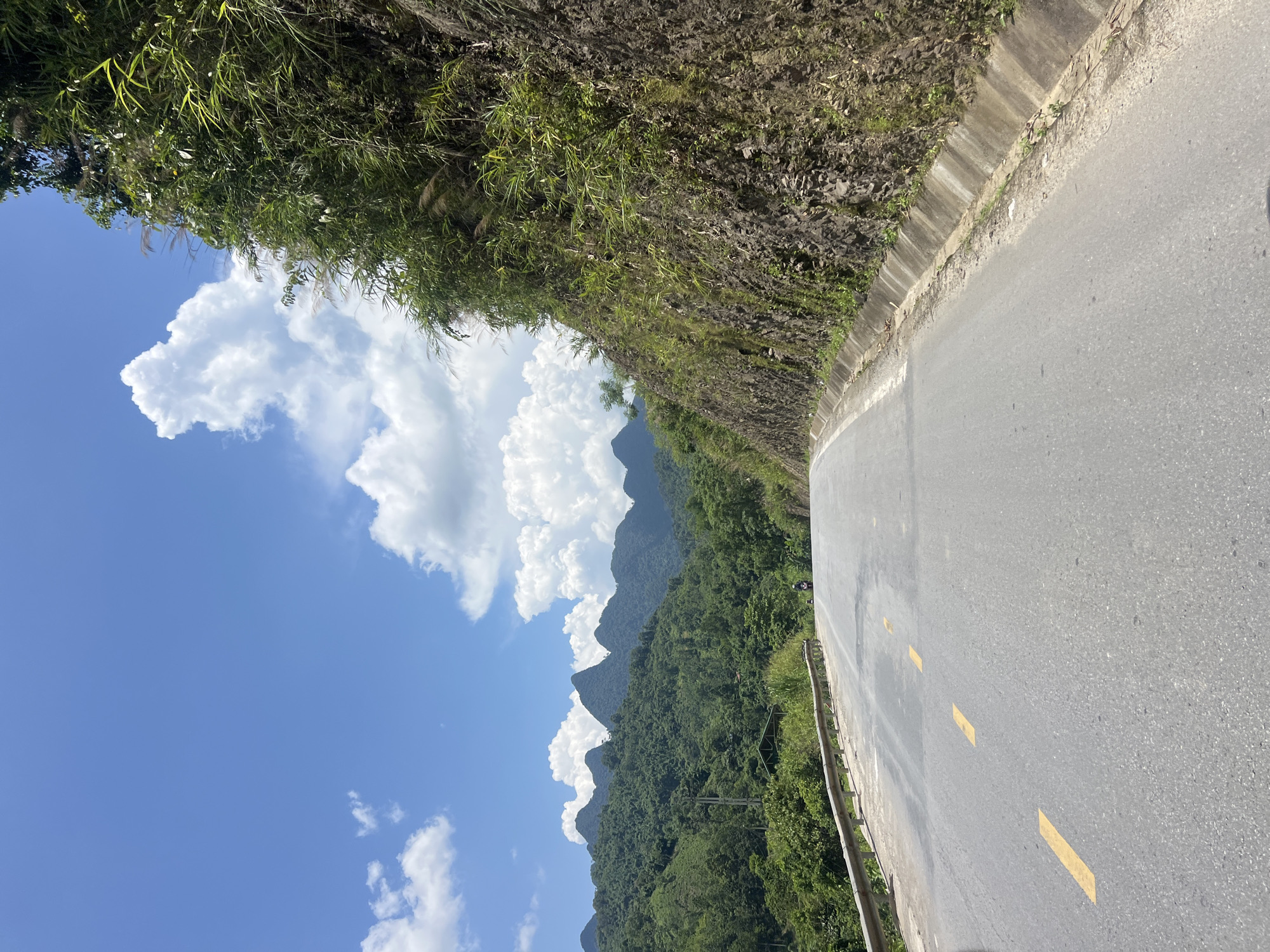 A paved road winds through a mountainous landscape under a bright blue sky with scattered white clouds. To the right, a steep, rocky embankment covered in lush green vegetation rises sharply, bordered by a concrete drainage channel. The road features a metal guardrail on the left side and yellow dashed lines marking the lanes. In the distance, a series of green, tree-covered mountains form the horizon, with a small building visible amidst the foliage on the left. A single motorcycle is present on the road, heading away from the camera. The scene depicts a scenic route in the rural environment of Minh Tân Commune, Vietnam.