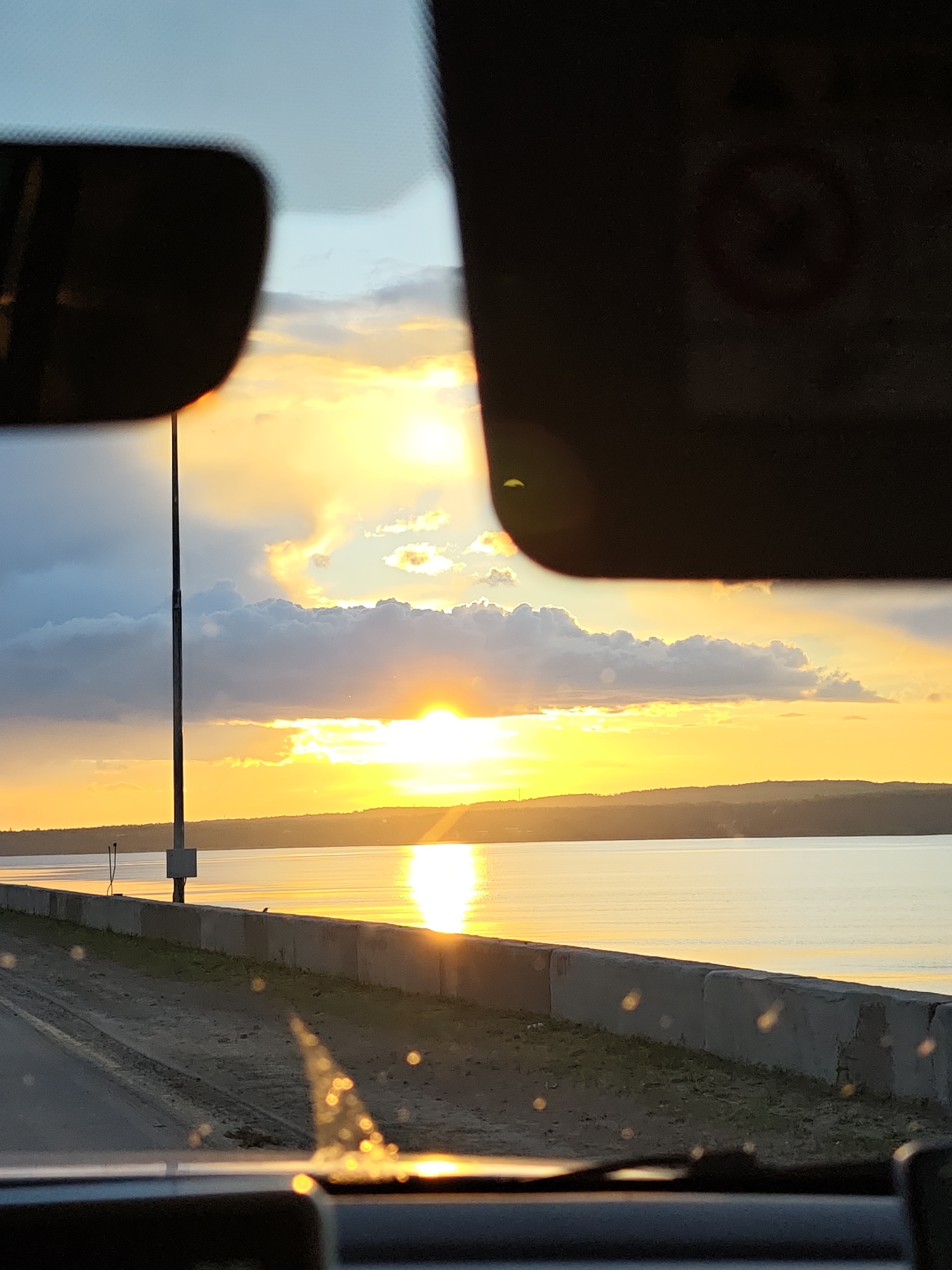The image shows a sunset view from inside a vehicle, likely a car, in Chaikovsky, Russia. The sky displays golden hues due to the setting sun, with clouds scattered around. A body of water, possibly the Kama River, reflects the sunlight. A concrete barrier runs along the edge of the road. Two sun visors on the vehicle's windshield frame the view. A utility pole stands on the roadside. The scene appears serene and peaceful, capturing a moment in time.
