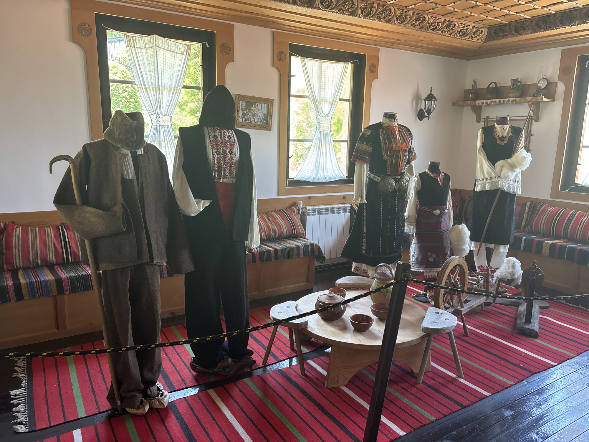 This image captures a detailed museum exhibit showcasing traditional Bulgarian attire and household items within a room designed to emulate a historical home, located in Belchin, Bulgaria. Five mannequins are arranged in a semicircle on a vibrantly striped red, green, black, and white woven rug that covers the central floor. A black rope barrier with yellow segments demarcates the display area.
From left to right, the mannequins display traditional clothing:
1. A male figure wears a dark brown, heavy, felt-like coat and trousers, a dark hat, and light-colored soft shoes, holding a curved wooden shepherd's crook.
2. A male figure wears a white shirt with red and black embroidered patterns, a black sleeveless vest, and red lower garments, topped with a dark head covering.
3. A female figure wears a dark dress with rich orange, black, and white embroidery on the bodice and sleeves, cinched with a wide, ornate silver belt.
4. A female figure wears a white embroidered shirt, a dark sleeveless vest, and a patterned skirt, adorned with a multi-strand necklace.
5. A female figure wears a white embroidered top, a dark tunic with white trim, and a patterned skirt, holding a wooden staff topped with raw white wool.
The room features white walls with carved wooden window frames and decorative circular motifs in the upper corners. Two large windows, draped with sheer patterned curtains, look out onto green foliage. Between the left windows, a framed black-and-white photograph is displayed. The ceiling is intricately crafted from carved wood with ornate floral and geometric patterns. Along the back wall, wooden benches topped with striped cushions provide seating. A white radiator is visible under the central window.
In the foreground, a low, round, light-colored wooden table holds several traditional ceramic pots, bowls, and a pitcher. Two small, three-legged wooden stools are positioned nearby. To the right of the table, a wooden spinning wheel and another wooden textile tool, possibly part of a loom, are displayed with balls of raw white wool. On the right wall, a rustic wooden shelf holds various ceramic vessels, a small woven basket, and an antique oil lamp. An ornate metal wall sconce with a lamp is mounted above the female mannequins. Dark wooden floorboards border the colorful rug.