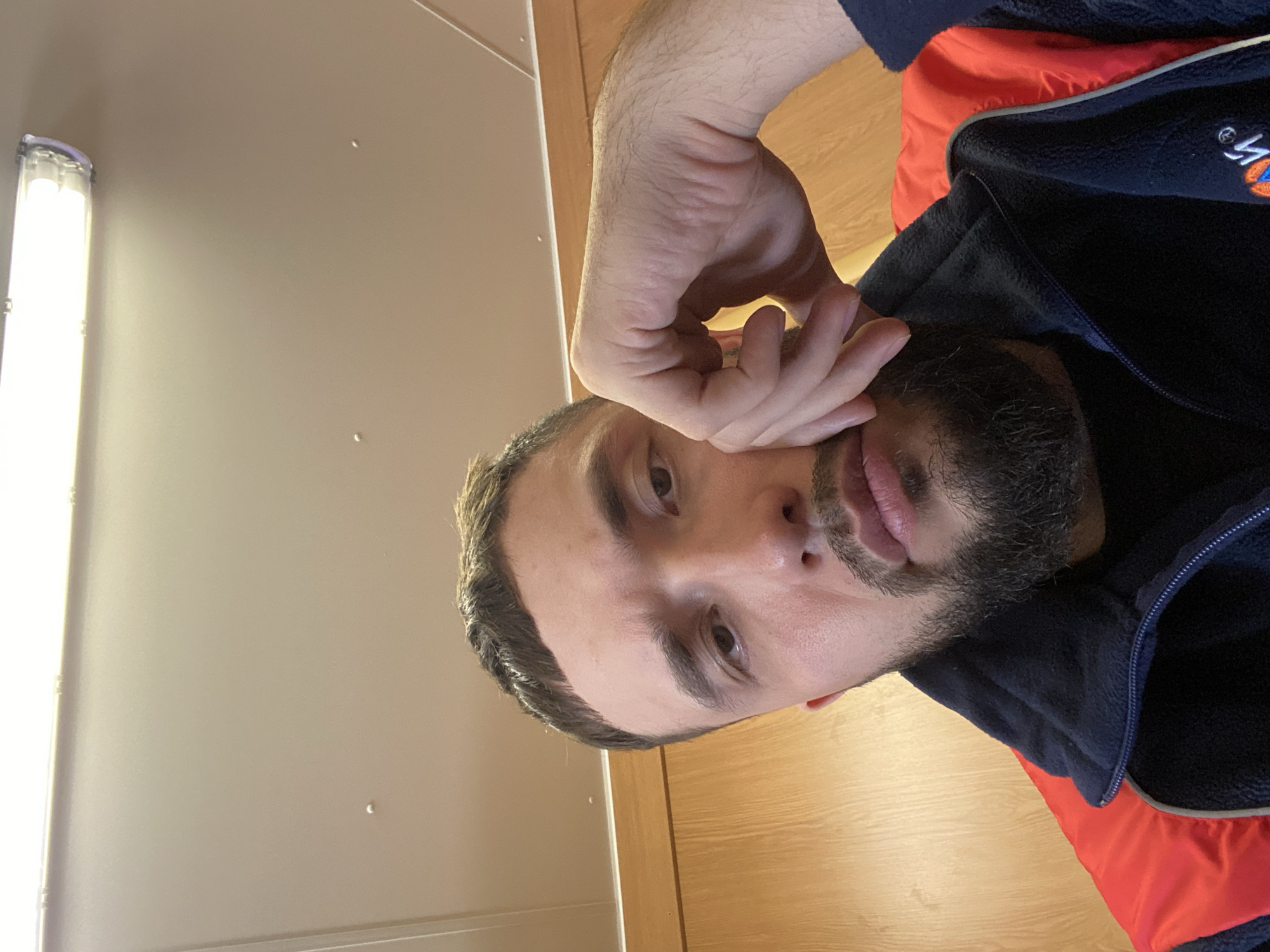 A selfie shows a man with a beard and dark hair. He is wearing a dark blue jacket with an orange lining. His hand is raised to his face. The background includes a beige wall with a fluorescent light fixture. It is not possible to determine from the image if it was taken in Luzhitsy, Russia.