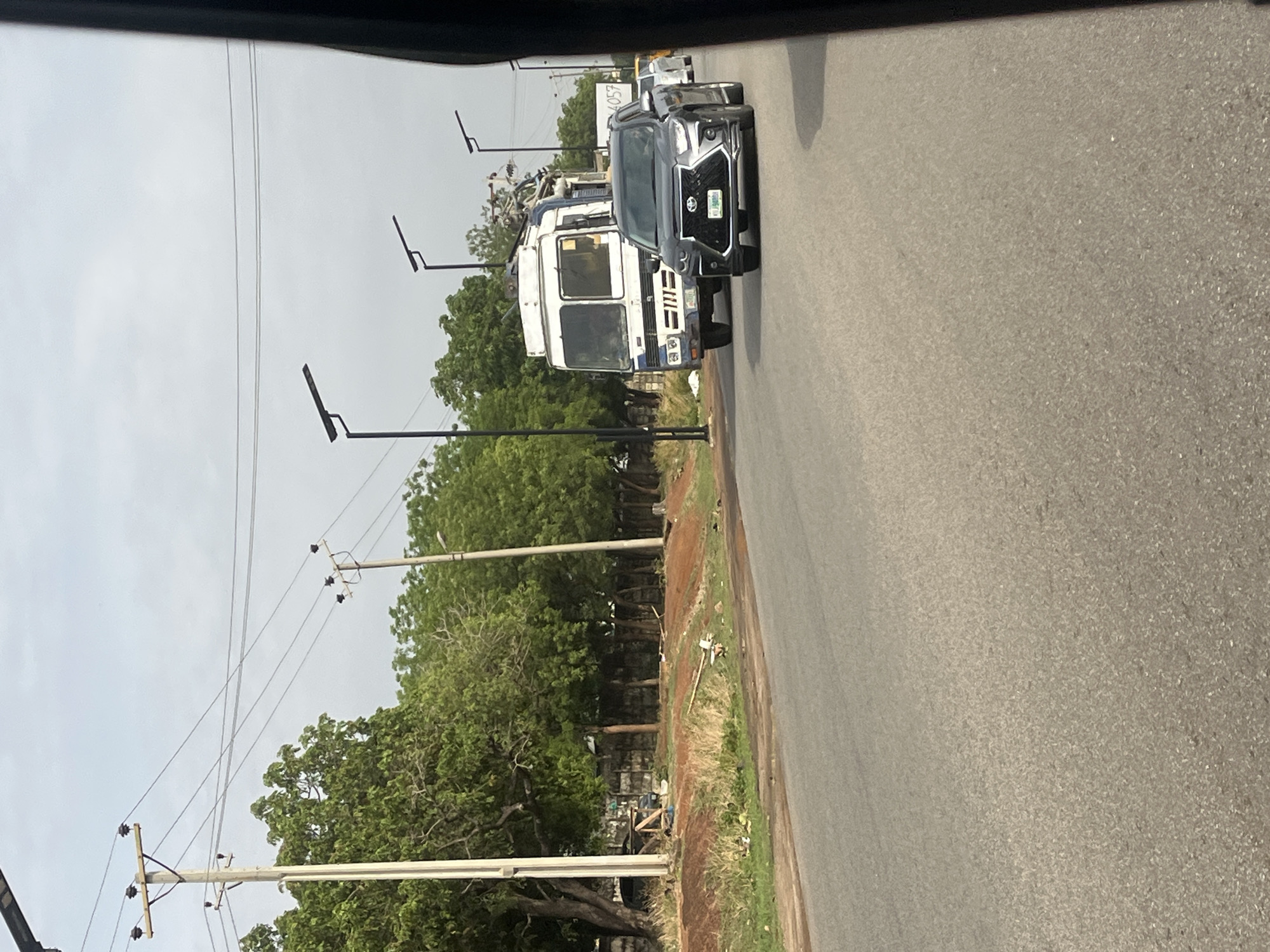 The image, taken from within a vehicle, depicts a road in Galadimawa, Nigeria. A black SUV is traveling on the right side of the road, followed by a white truck loaded with various objects. The road is paved and appears to be in fair condition. On the left side of the road are trees and utility poles. The sky is overcast. No people are clearly visible.
