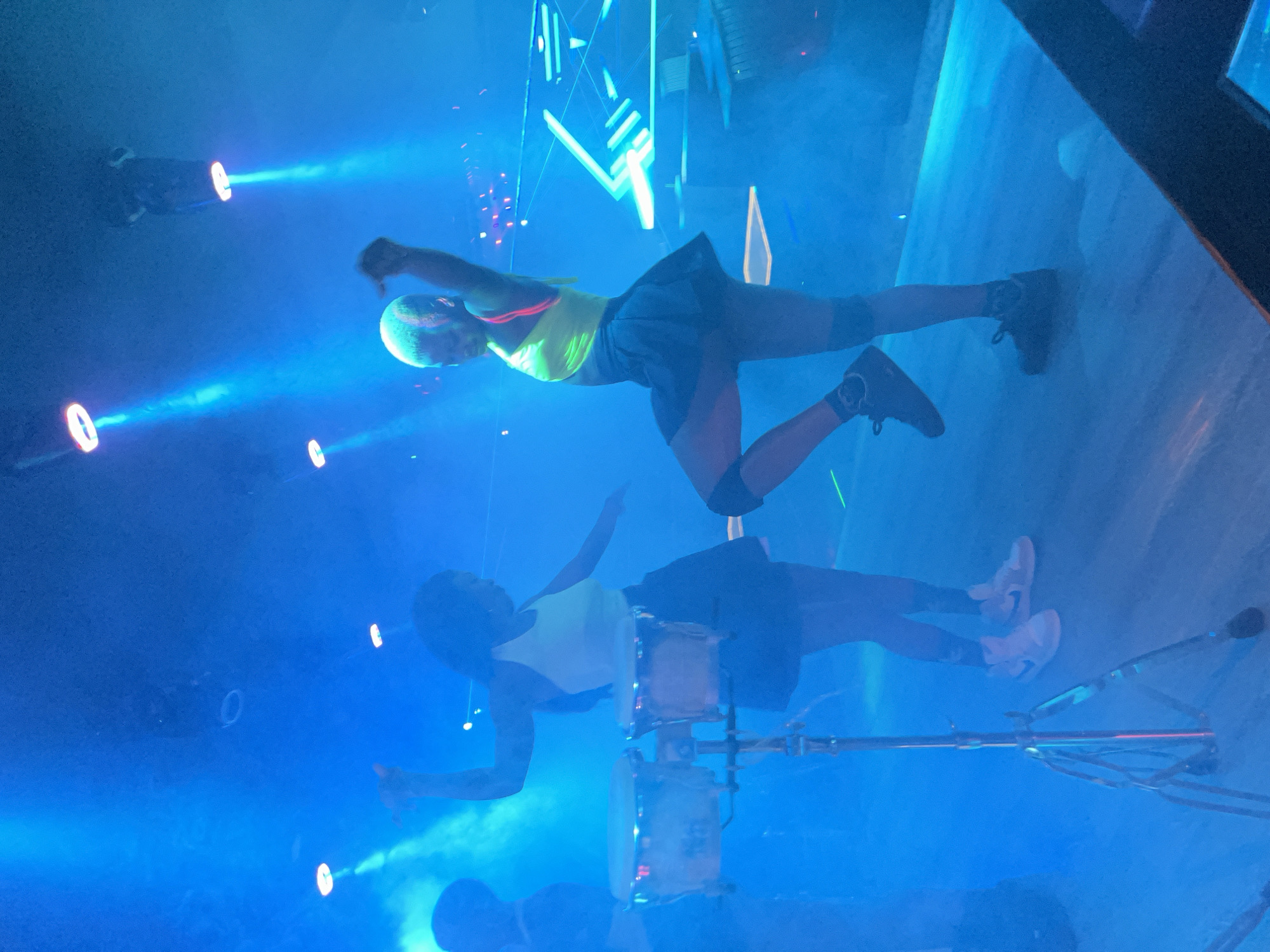 Two women are performing on a stage in a dimly lit venue. One woman, on the left, wears a light-colored top and a dark skirt, and stands with her arms raised. The other woman, on the right, wears a neon yellow top and a dark skirt, and is captured mid-dance with her left leg extended. A drum set is positioned in the foreground on the left side of the image. The background features blue stage lighting and fog effects, with abstract geometric light installations visible on the right. The overall atmosphere suggests a musical or dance performance. The location is described as being in the city of Howayi, Nigeria.