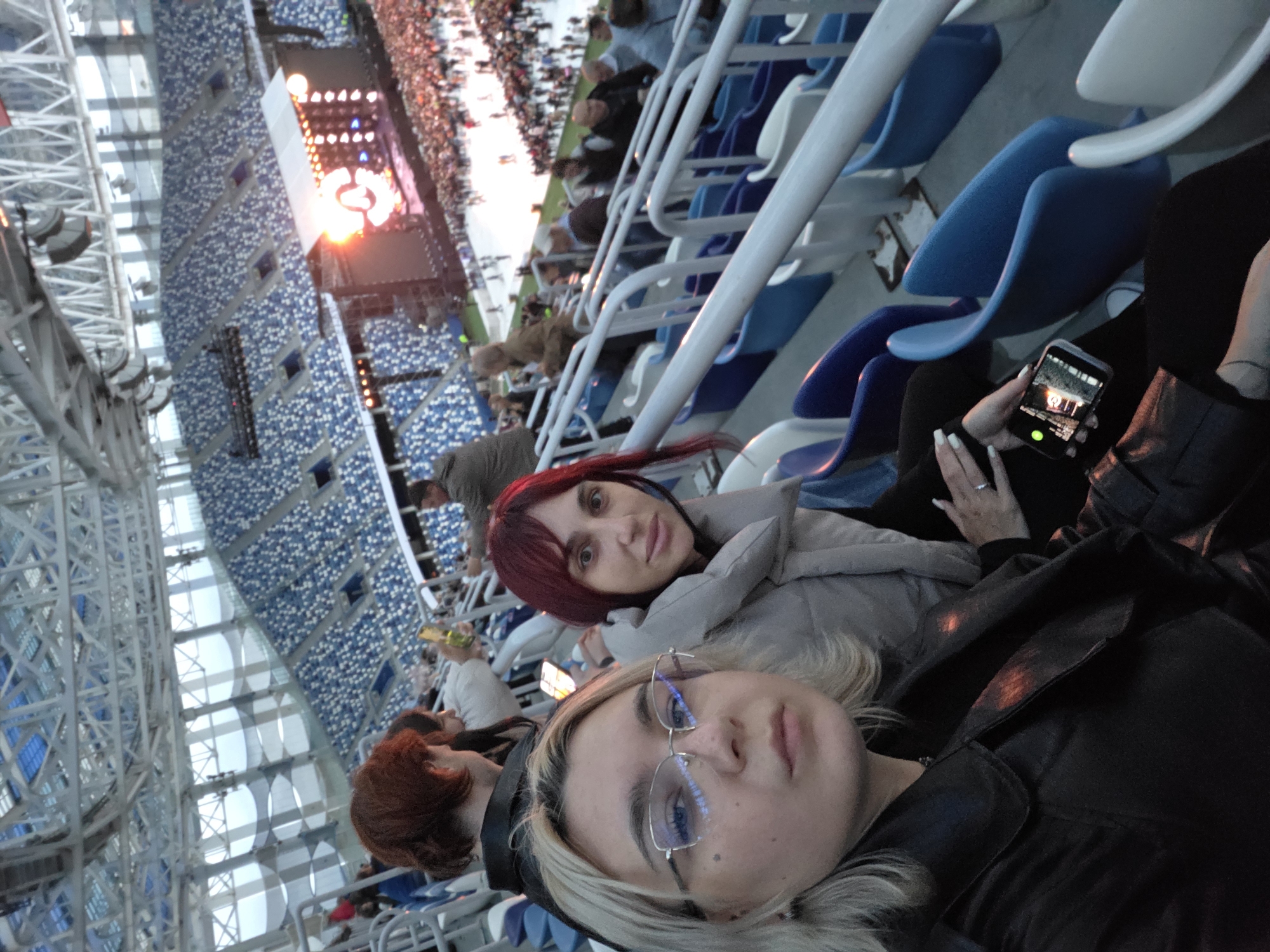 The image shows a group of people in a stadium, likely at a concert or sporting event, in Nizhny Novgorod, Russia. The stadium has tiered seating in blue and white, with a large stage and screen visible in the background. The lighting suggests it is either evening or overcast. Two women are in the foreground, with one taking a selfie. Both are dressed casually. The woman on the left is wearing glasses and a black jacket, while the woman on the right has red hair and a light-colored puffy jacket. Other spectators are visible in the background, some holding up their phones. The overall scene depicts a lively event attended by a diverse crowd.
