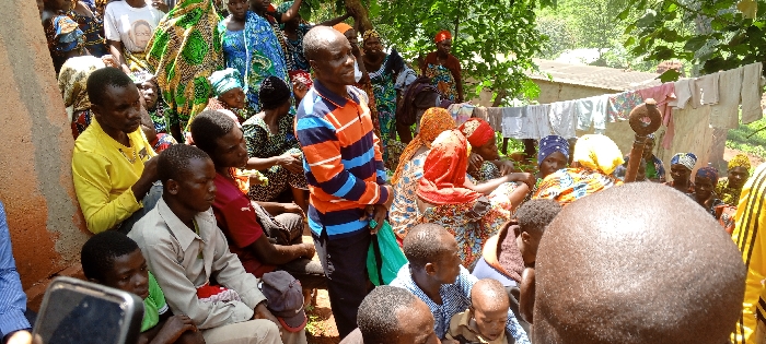 A group of people are gathered outside, seemingly in a rural or less developed area. Most are dark-skinned and are wearing casual clothing, some in brightly patterned fabrics. There are men and women of varying ages. A man stands out in the middle, wearing an orange and blue striped shirt. The group appears to be sitting or gathered around something out of frame, possibly a discussion or event. Laundry hangs on a line in the background near a simple structure.