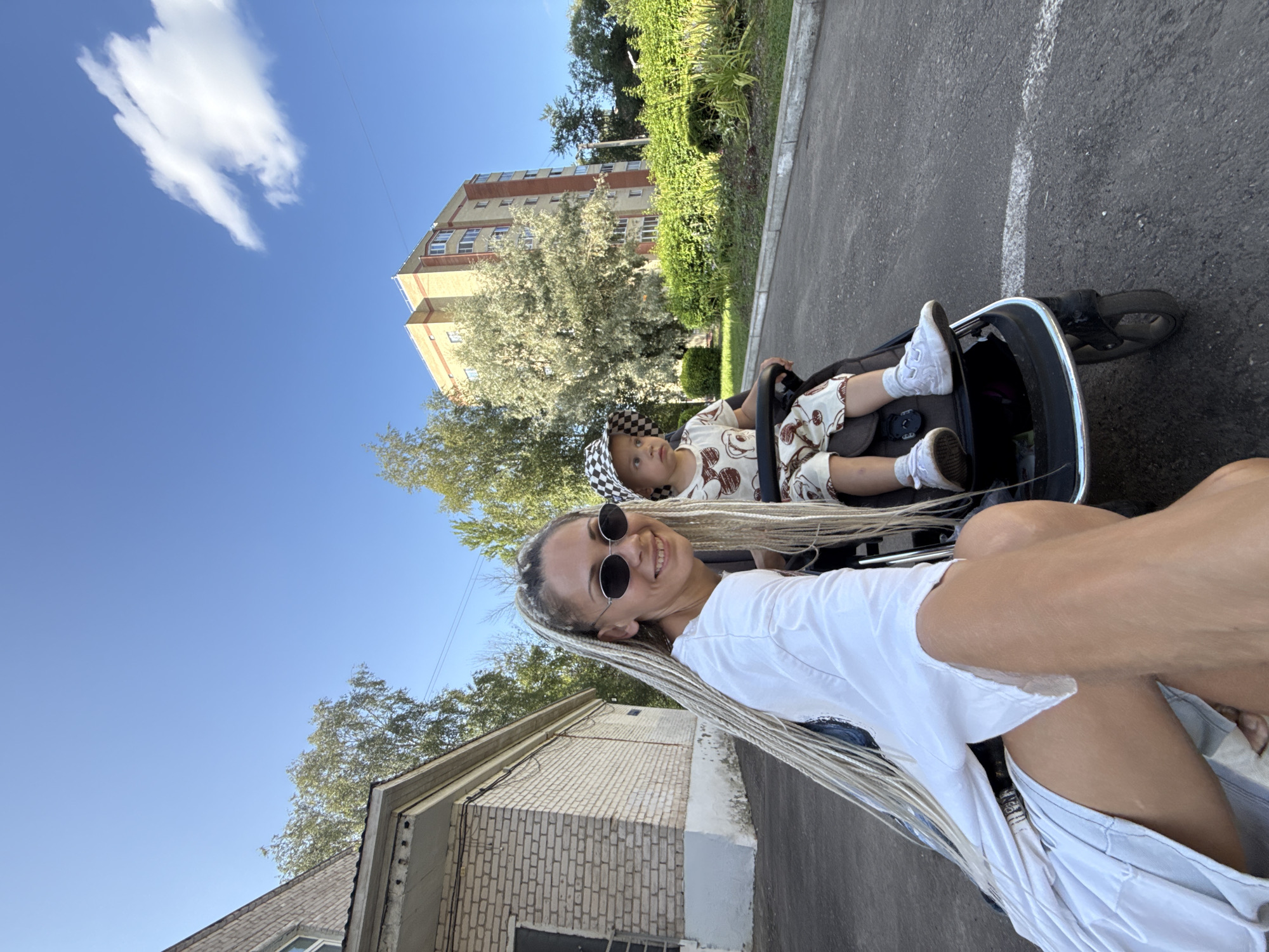 A woman with long blonde braids and sunglasses is pushing a stroller with a child in it. The woman is wearing a white t-shirt and a light blue skirt. The child is wearing a patterned outfit and a checkered hat. They are on a paved path next to a brick building and a residential building. There are trees and bushes in the background. The sky is blue with a few clouds. The location is Rusanovo, Russia.