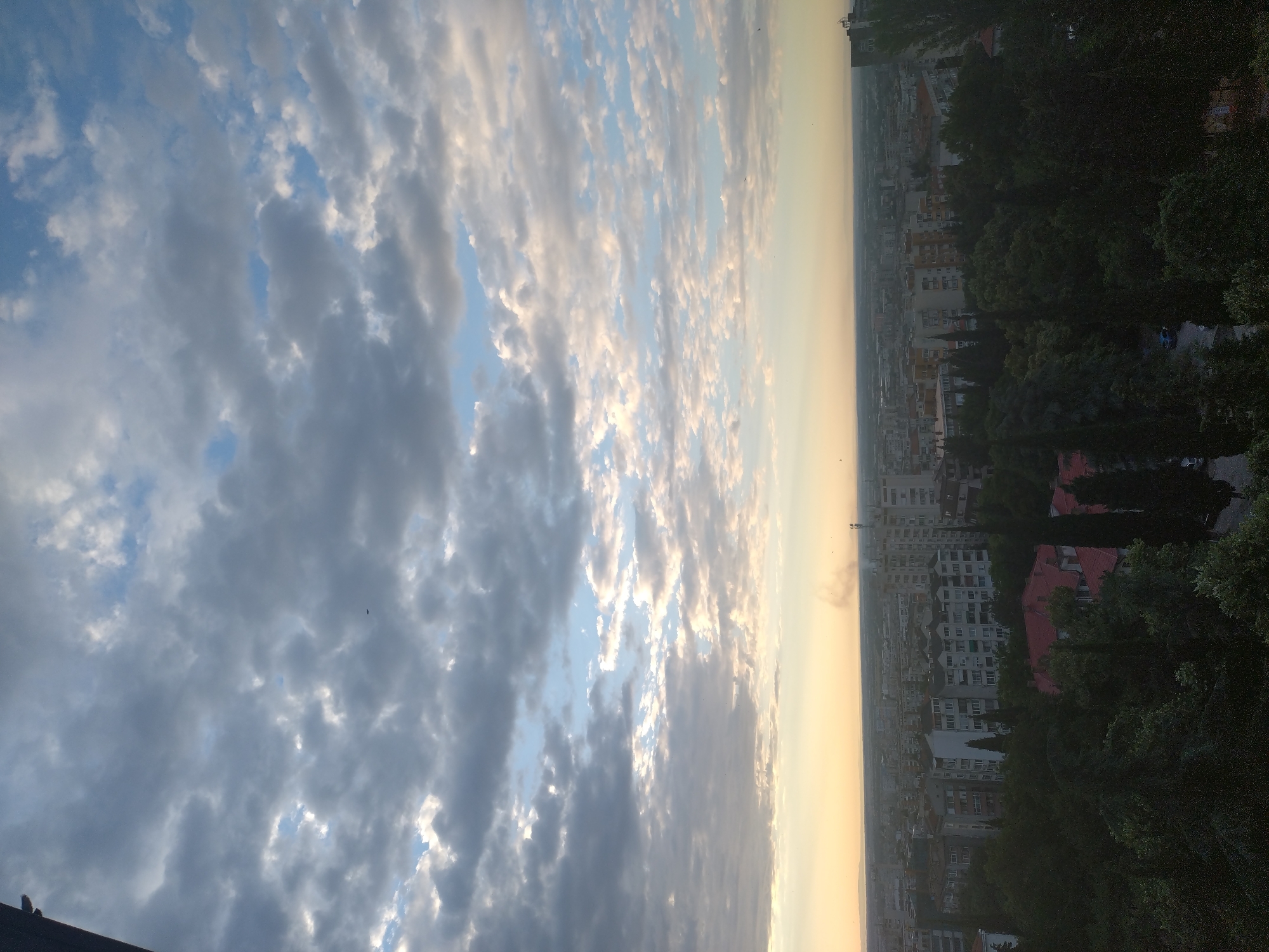 This is a high-angle, vertical, full shot of a cityscape at dusk. The sky is filled with clouds, ranging from dark grey to light grey and white, with patches of blue sky visible. The sun is low on the horizon, casting a warm, yellow-orange glow. Below the sky is a dense urban landscape, primarily composed of apartment buildings of various heights and designs, with some flat roofs and others with red tile. Lush green trees, some appearing to be cypress trees, form a dense canopy in the foreground and between buildings. There is a plume of smoke rising from the right side of the city. The scene appears to be a view of the city of Stara Zagora, Bulgaria. No people are visible in the image.
