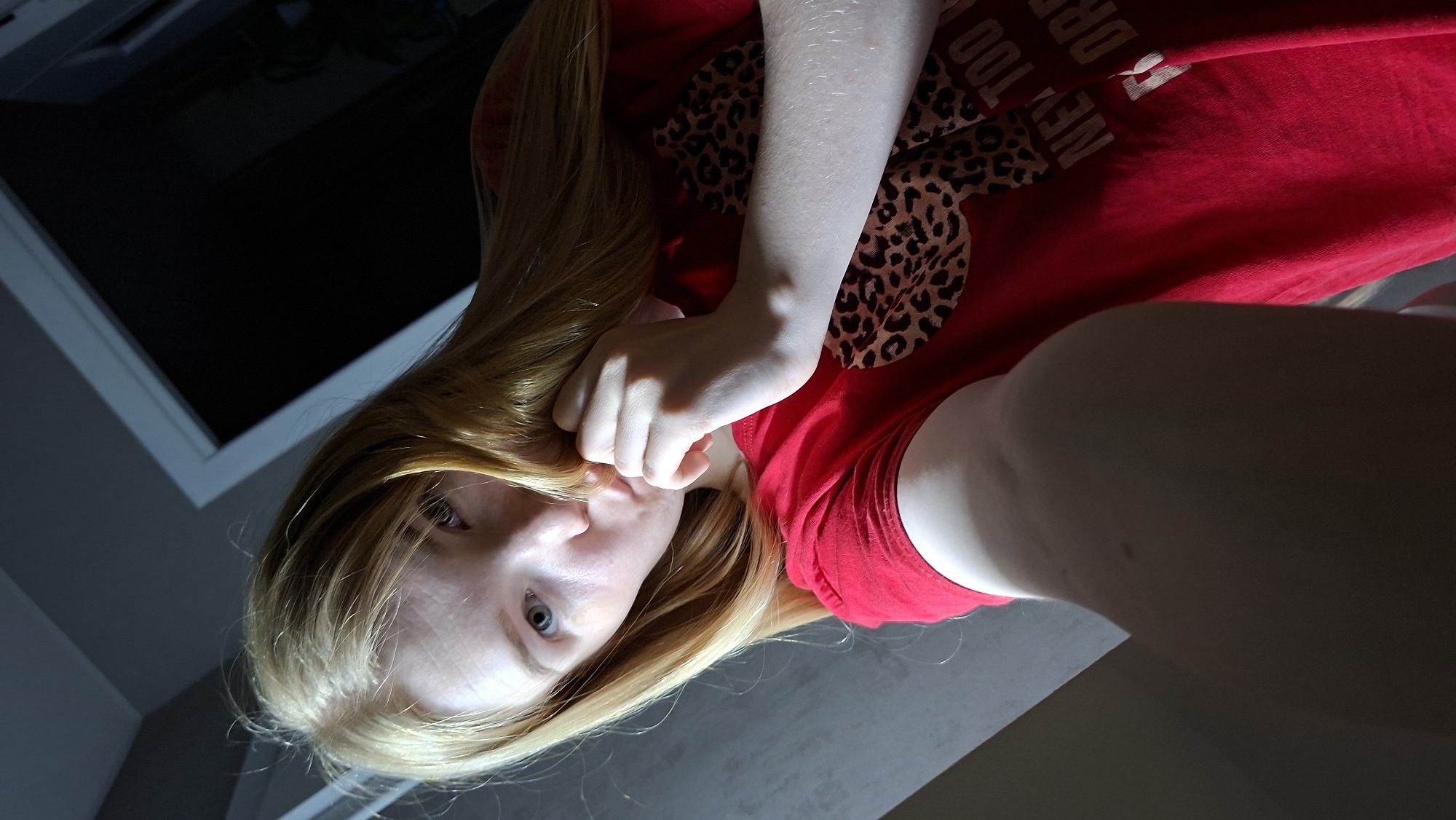A low-angle selfie depicts a fair-skinned person with blonde hair and blue eyes. The person is wearing a red t-shirt with a leopard print design. The background includes a dark area, possibly a room, and a window frame. The location, Chaikovsky, Russia is not discernible.