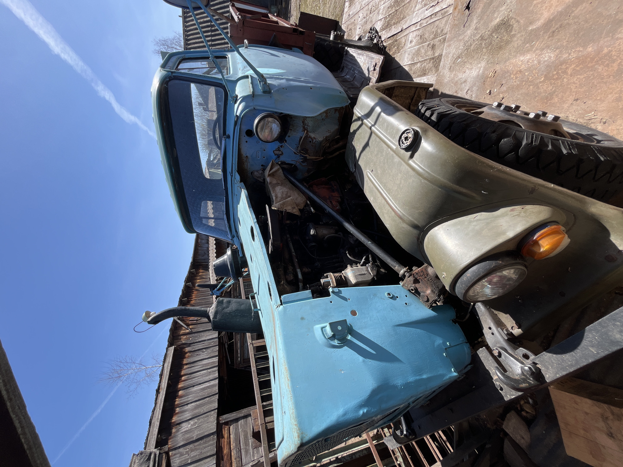 The image shows a dilapidated, light blue and olive green truck with missing body panels. The truck is situated in what appears to be an outdoor storage or salvage yard, with an old wooden building visible in the background. The sky is clear with a contrail. I do not have information about the specific geographic location of the photo.