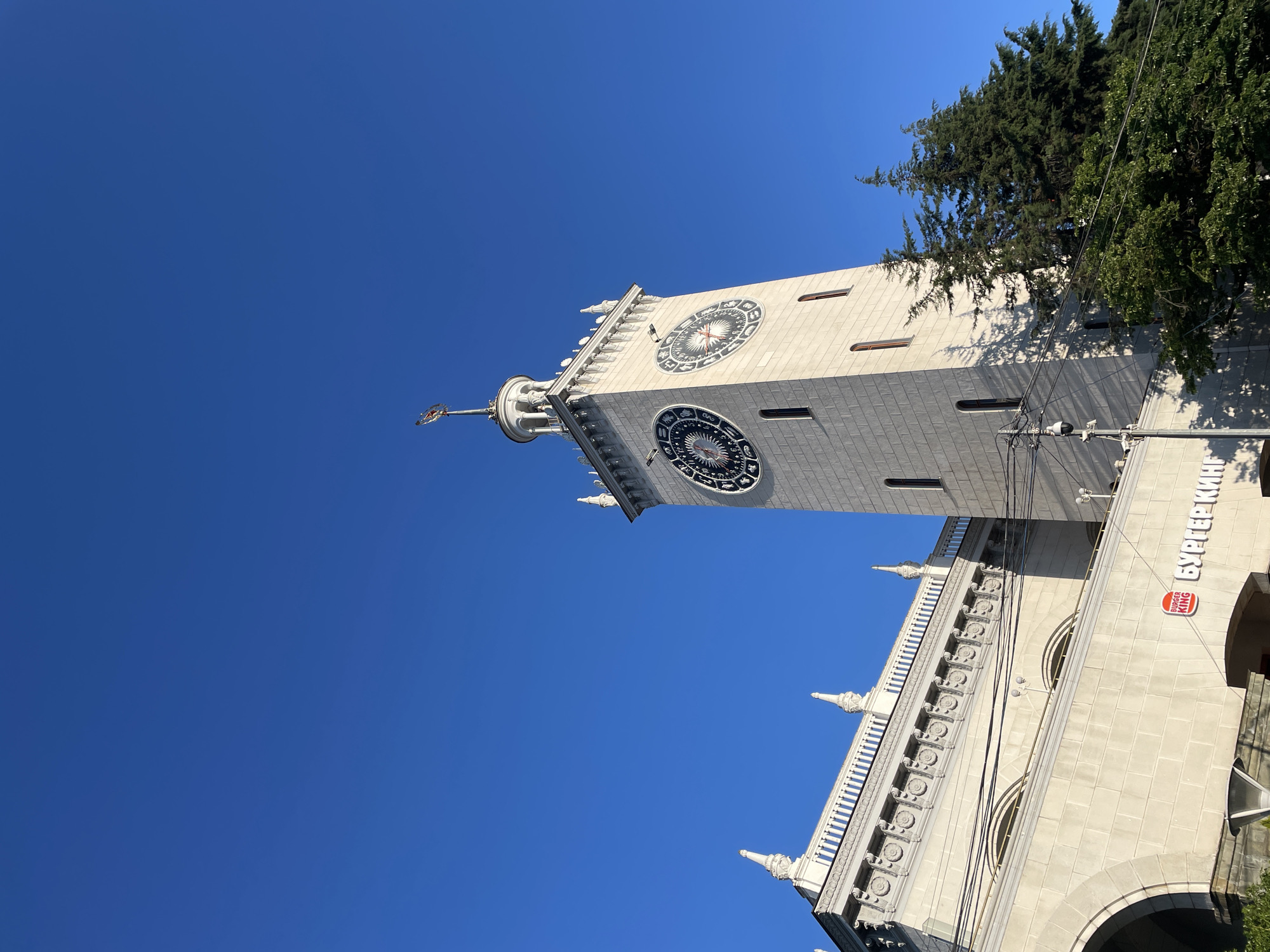 The image displays the iconic clock tower of the Sochi Railway Station, a prominent landmark in Sochi, Russia. The tower, constructed of light-colored stone, features large, ornate clock faces with zodiac symbols. Adjacent to the tower is a white building with classical architectural elements, including decorative cornices and pointed finials. A Burger King sign is visible on the lower portion of the white building. Evergreen trees, casting long shadows from the sun, are situated to the right of the clock tower. The sky above is a clear, vibrant blue. The photograph captures the station from a low angle, emphasizing the height of the tower and the grandeur of the architecture.
