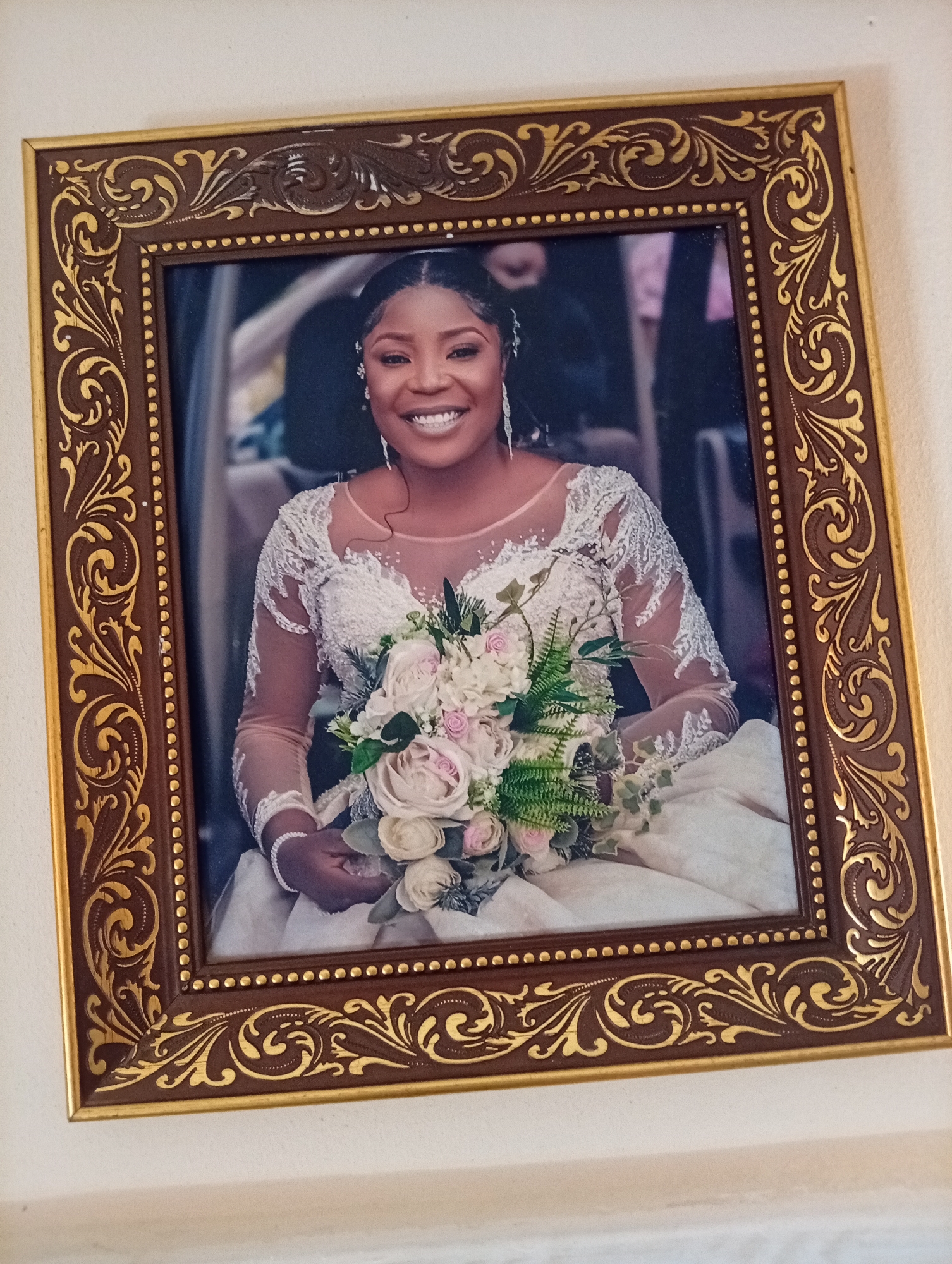 A framed portrait of a smiling bride in a wedding dress, holding a bouquet of flowers, is displayed on a wall. The bride has dark skin, dark hair styled in braids, and is wearing jewelry. The wedding dress is white with lace detailing. The bouquet consists of white and pink roses, along with greenery. The frame is ornate, with a dark brown wood finish and gold detailing. The photograph is positioned on a light-colored wall, suggesting it is likely within a home or a venue. No specific location within Gura Top, Nigeria, can be determined from the image.