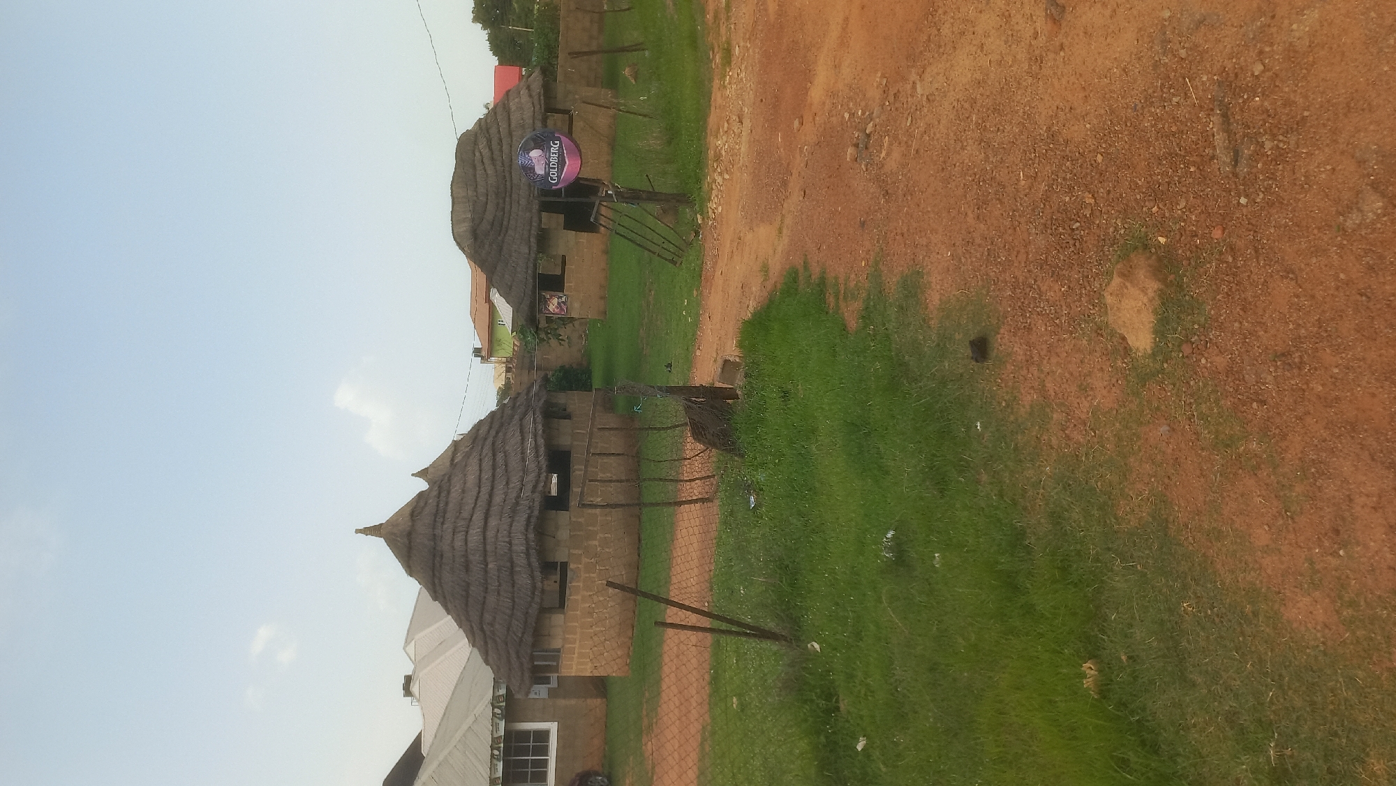 This media file captures an outdoor scene in Gura Top, Nigeria. The image features two thatched-roof buildings with brick walls, characteristic of traditional or rustic architecture. In front of the buildings, there is a well-maintained green lawn and a dirt path. A wire fence separates the lawn from the path and the buildings.
On the left side of the image, a modern building with white siding and windows is visible, suggesting a mix of old and new structures in the area. A circular sign with the word "GOLDBERG" in large letters and a logo is prominently displayed, likely indicating a commercial establishment.
The sky is clear and appears to be daylight, with some faint clouds. The overall atmosphere of the scene is calm and serene, with no people or specific activities depicted. The sunlight casts shadows on the grass and dirt path, indicating it's likely daytime. The presence of traditional structures alongside modern ones hints at a developing or transitional setting.
