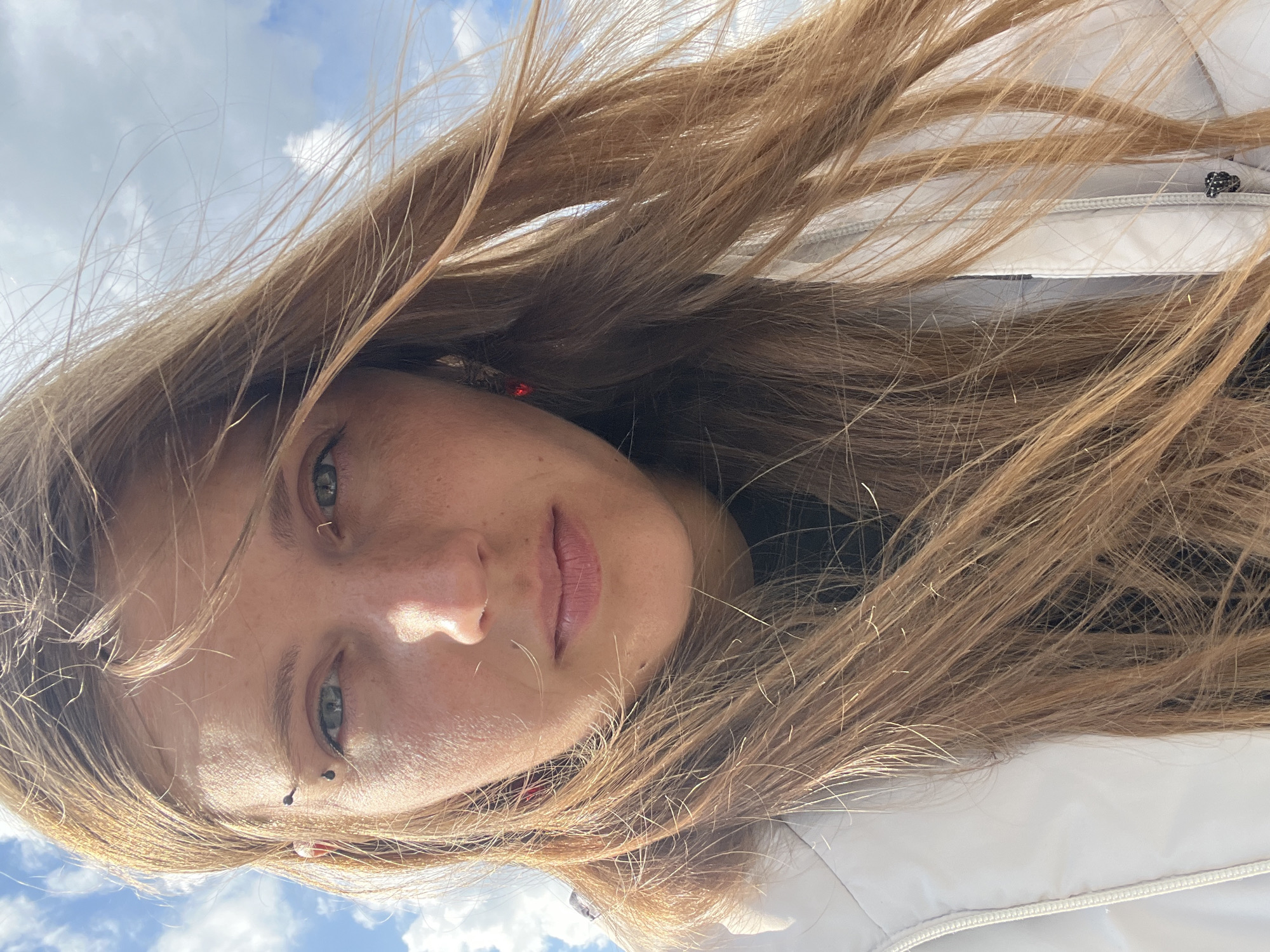 A close-up, frontal, waist-up, selfie-style photograph shows a woman with long, light brown, wind-blown hair and fair skin. She has green eyes, light freckles across her nose and cheeks, and is wearing a light-colored jacket with a zipper. She has a eyebrow piercing on her left eyebrow and small red earrings. The background is a bright blue sky with scattered white clouds. The image appears to have been taken outdoors on a sunny day. The image quality is sharp with natural lighting. The exact location in the city of Minsk, Belarus is not determinable from the visual information provided.
