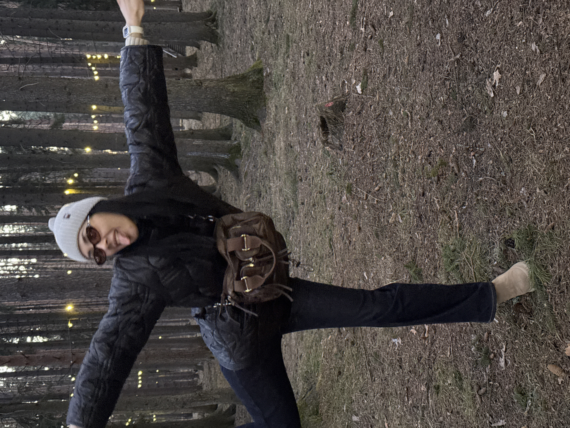 The image portrays a person striking a dynamic, playful pose in a forest setting, likely in Votkinsk, Russia. The individual, an adult with dark hair, is dressed for cool weather, wearing a grey knit beanie, sunglasses, a dark puffer jacket, dark pants, and light-colored boots. They extend both arms horizontally and one leg to the side, balancing or dancing. Their tongue is slightly out, conveying a lively and cheerful expression.
A brown, structured backpack-style purse with metallic buckles and zippers is worn across their body, and a light-colored digital watch is visible on their left wrist. The background features numerous slender, dark tree trunks, indicative of a coniferous forest. Small, warm-toned string lights are visible strung between the trees, suggesting it is either dusk or early evening, or an artificially illuminated outdoor space. The ground is covered with dry, sparse grass, fallen leaves, and pine needles, typical of late autumn, winter, or early spring. No visible text is present in the image.