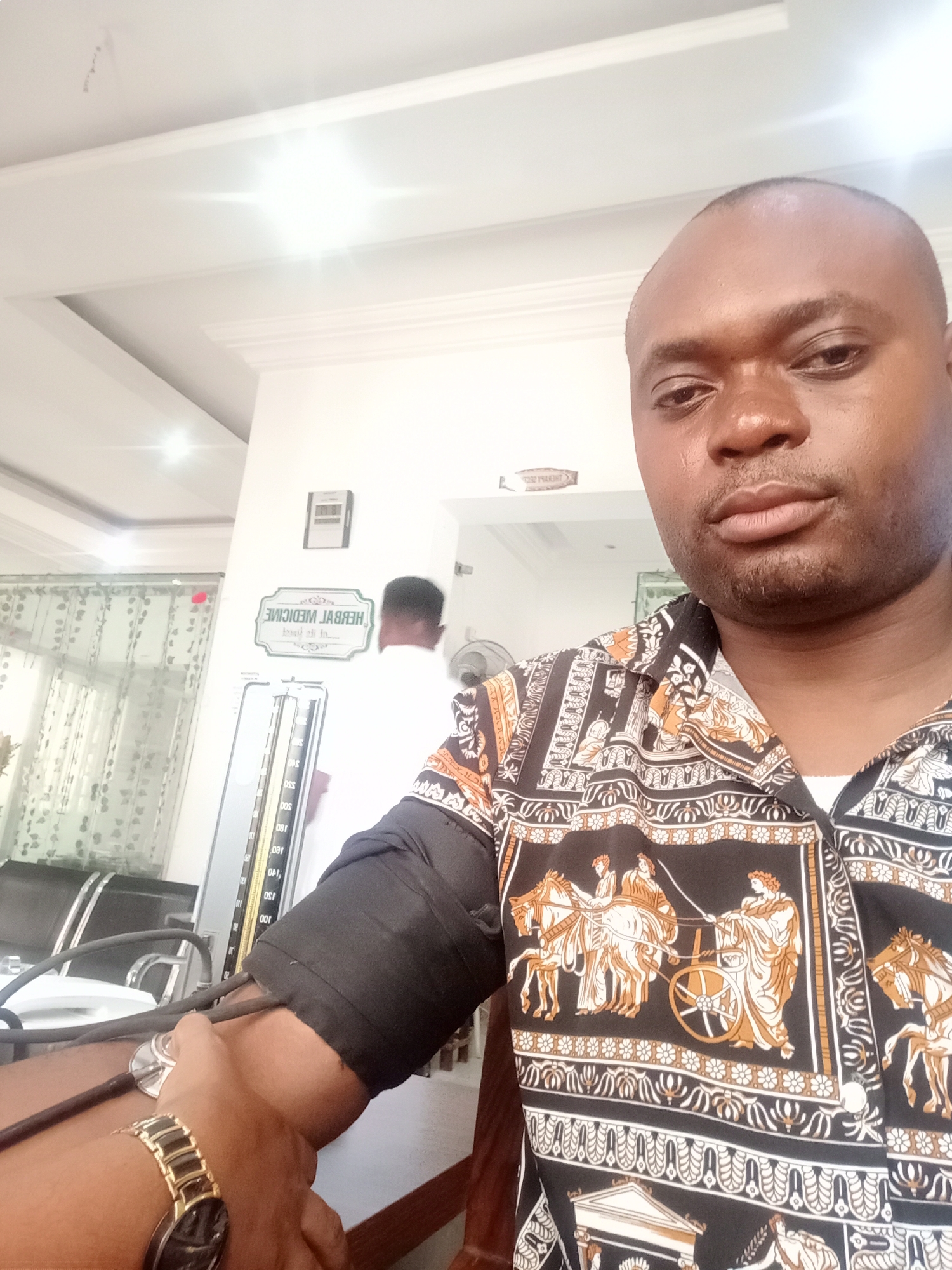 The image is a selfie taken by a dark-skinned man in what appears to be a medical setting in Gwarinpa, Nigeria. The man is wearing a patterned button-down shirt with black and gold designs, and a gold wristwatch on his left wrist. A blood pressure cuff is fastened around his upper left arm, connected to a sphygmomanometer standing on a table. Behind him, another person in a white coat is partially visible. A sign that reads "HERBAL MEDICINE" is visible on the wall, written in reverse. A digital clock is mounted above the sign, and a sign is posted above a doorway that reads "TOILET". The room has white walls and ceiling, and a partially visible window with patterned curtains. The setting suggests a medical clinic or doctor's office.