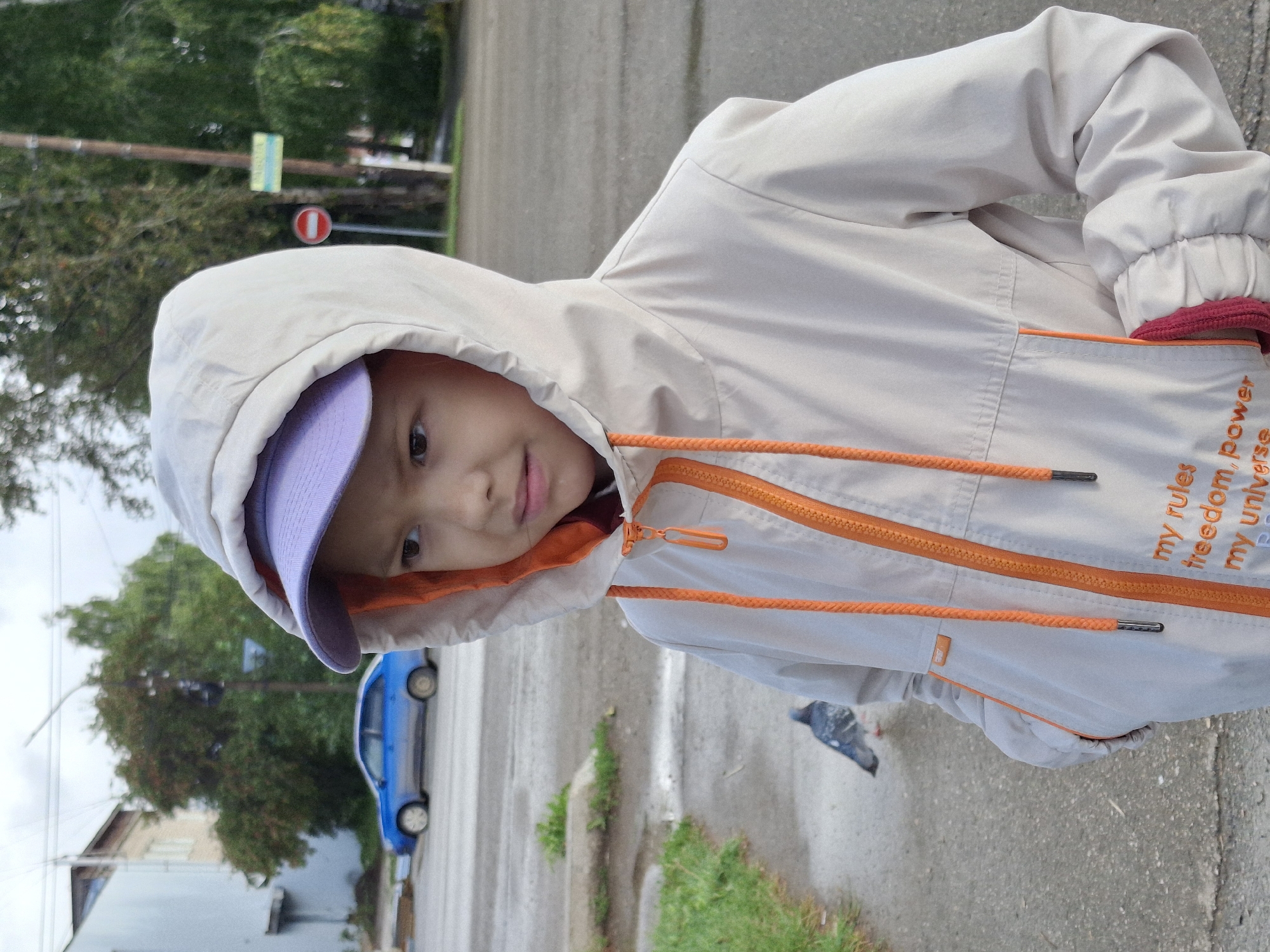 A medium shot captures a young person in a hooded, cream-colored jacket with orange trim and drawstrings, standing outdoors in what appears to be Elovo, Russia. The person's head is turned to the right, with their face visible in profile. A light purple baseball cap is worn under the hood. In the background, a blue car is parked on a wet street. To the left, a pigeon stands on a wet sidewalk. The environment suggests a cloudy, potentially damp day with trees and a building visible. A red and white circular traffic sign is also present in the background. The jacket has text printed on the lower front, partially visible.