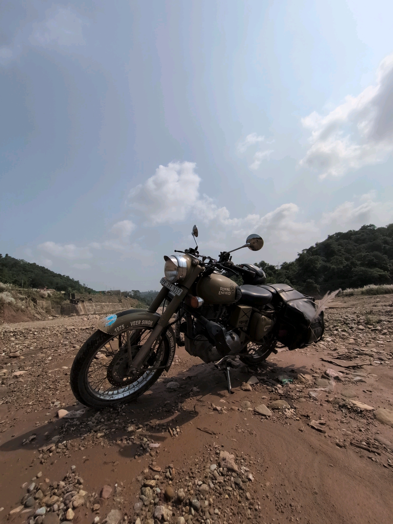 A tan Royal Enfield motorcycle is parked on a rocky, dirt path with scattered stones and patches of mud. The motorcycle is angled towards the right side of the frame and is equipped with saddlebags. The background features a dry riverbed with a concrete structure, hills covered in lush green trees, and a bright, cloudy sky. Text on the motorcycle's fender reads "VEER SINGH 1975". The scene is set in Jaanta, India, specifically on a rocky, semi-arid terrain adjacent to a water body or dam.