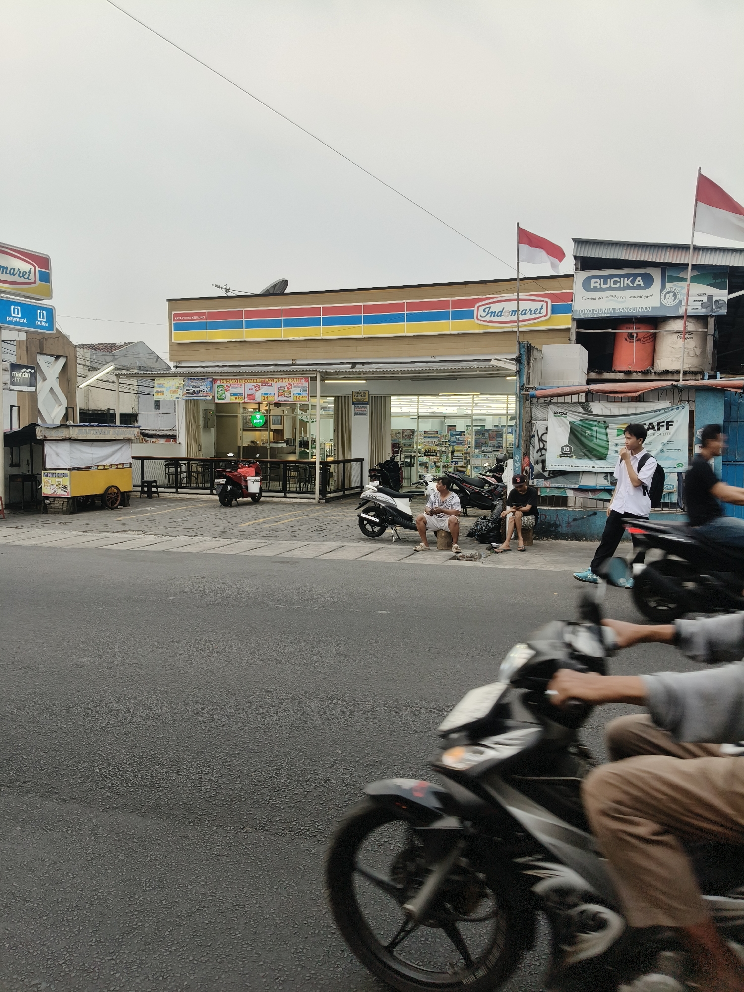 The image depicts a street scene in front of an Indomaret convenience store, likely located in South Tangerang, Indonesia, given the presence of Indonesian flags. The store, characterized by its distinctive red, yellow, and blue striped facade, is situated along a paved road. To the left of the store, a "mandiri atm" sign is visible, along with a yellow food cart covered with a white tarp.
Several individuals are present in the scene. Two men are seated on low stools outside the store; one is wearing a patterned t-shirt and shorts, and the other is wearing a dark t-shirt and shorts. A young man in a white shirt, black pants, and a backpack stands near the entrance of an adjacent building, looking towards the left. To the right, two men are riding motorcycles, with one in the foreground blurred by motion. There are also several motorcycles parked in front of the store.
The environment includes the street with asphalt paving, sidewalks, and the buildings lining the road. Two Indonesian flags are prominently displayed, one on a pole to the left of the Indomaret sign and another on a building to the right. The sky is overcast. The scene captures a typical urban streetscape with a focus on commercial activity and pedestrian and vehicular traffic.